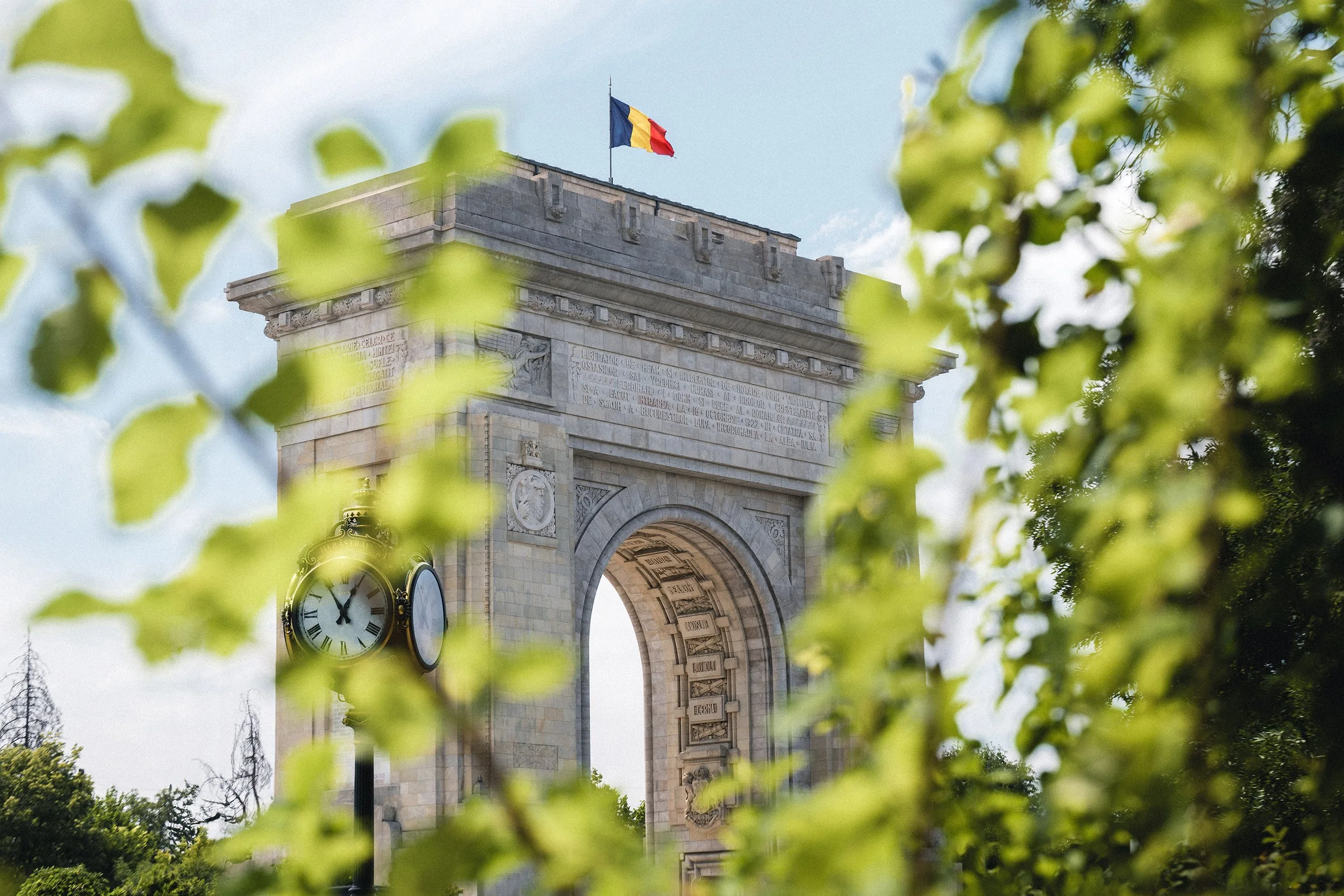 The Arc de Triomphe in Romania with a Romanian flag on top, viewed through green leaves.