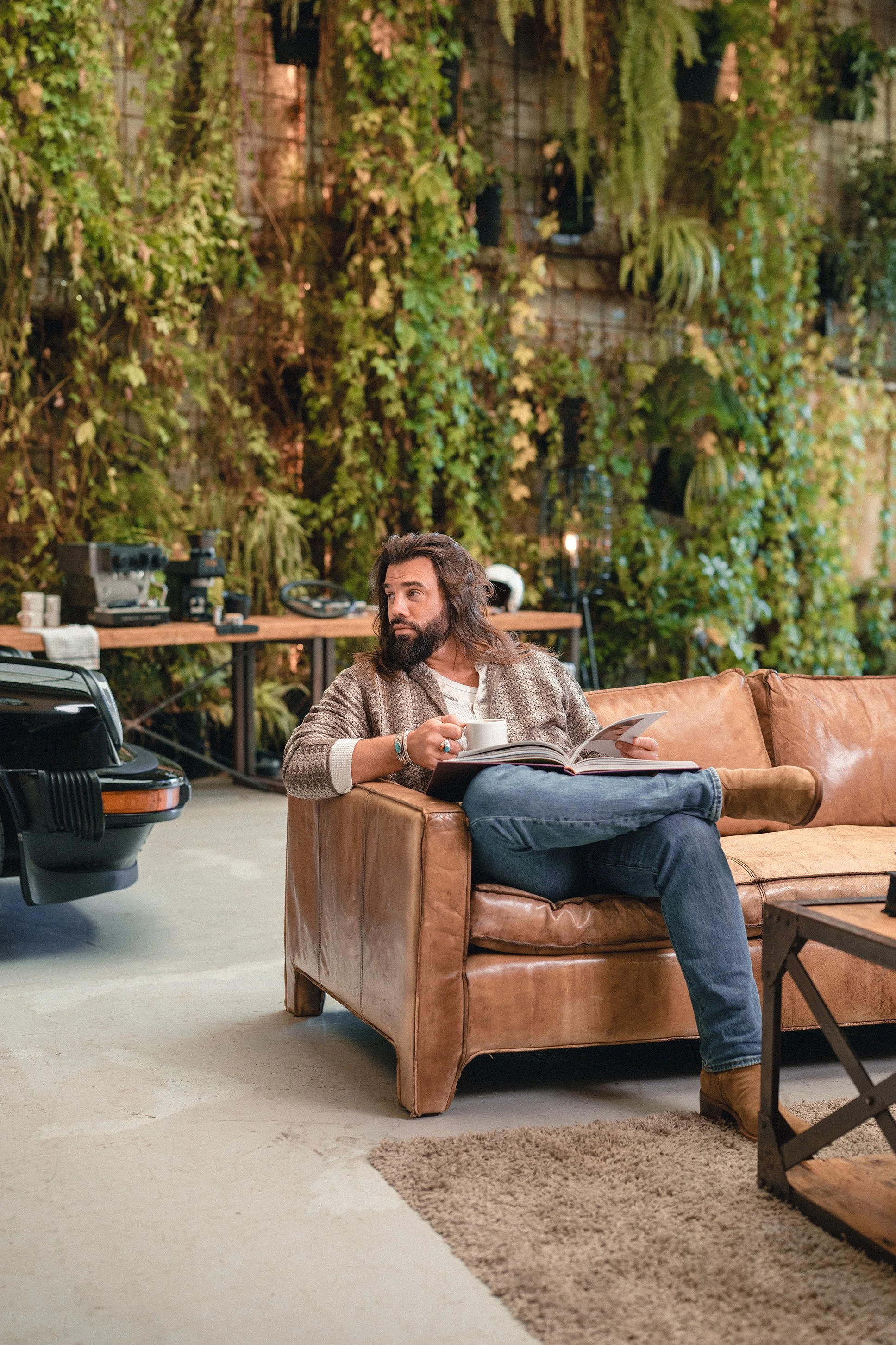 A man with long hair and a beard sitting on a tan leather sofa, holding a cup and reading a magazine in a cozy, green indoor space.