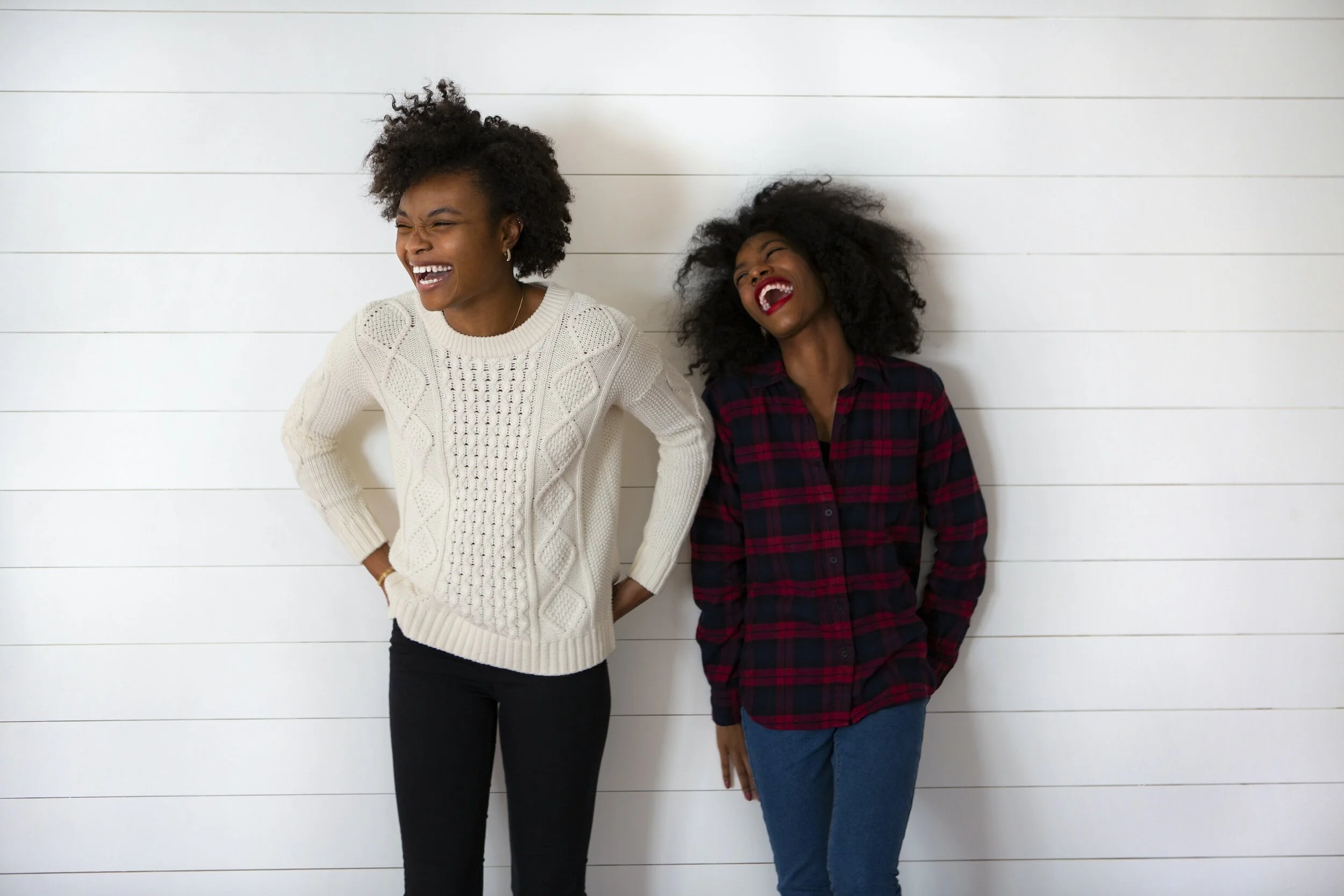 Two women with curly hair laughing and smiling while standing against a white wooden wall.
