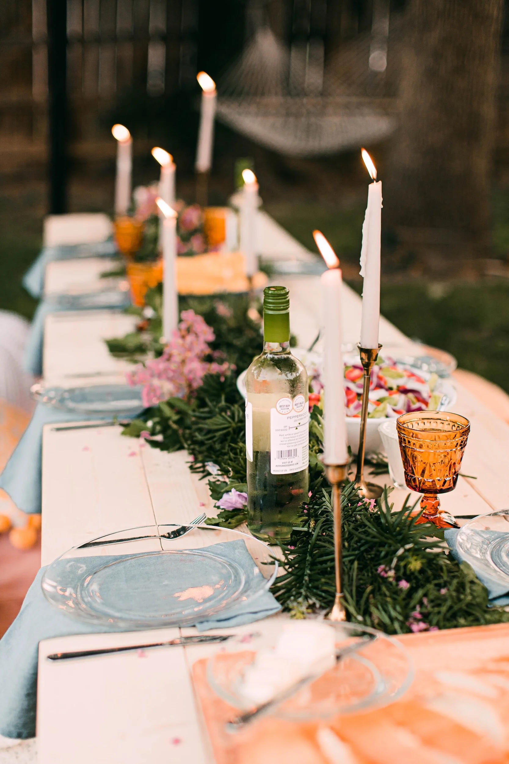 A decorated outdoor dining table set for a meal with plates, utensils, and glassware, lit candles, greenery, and pink flowers.