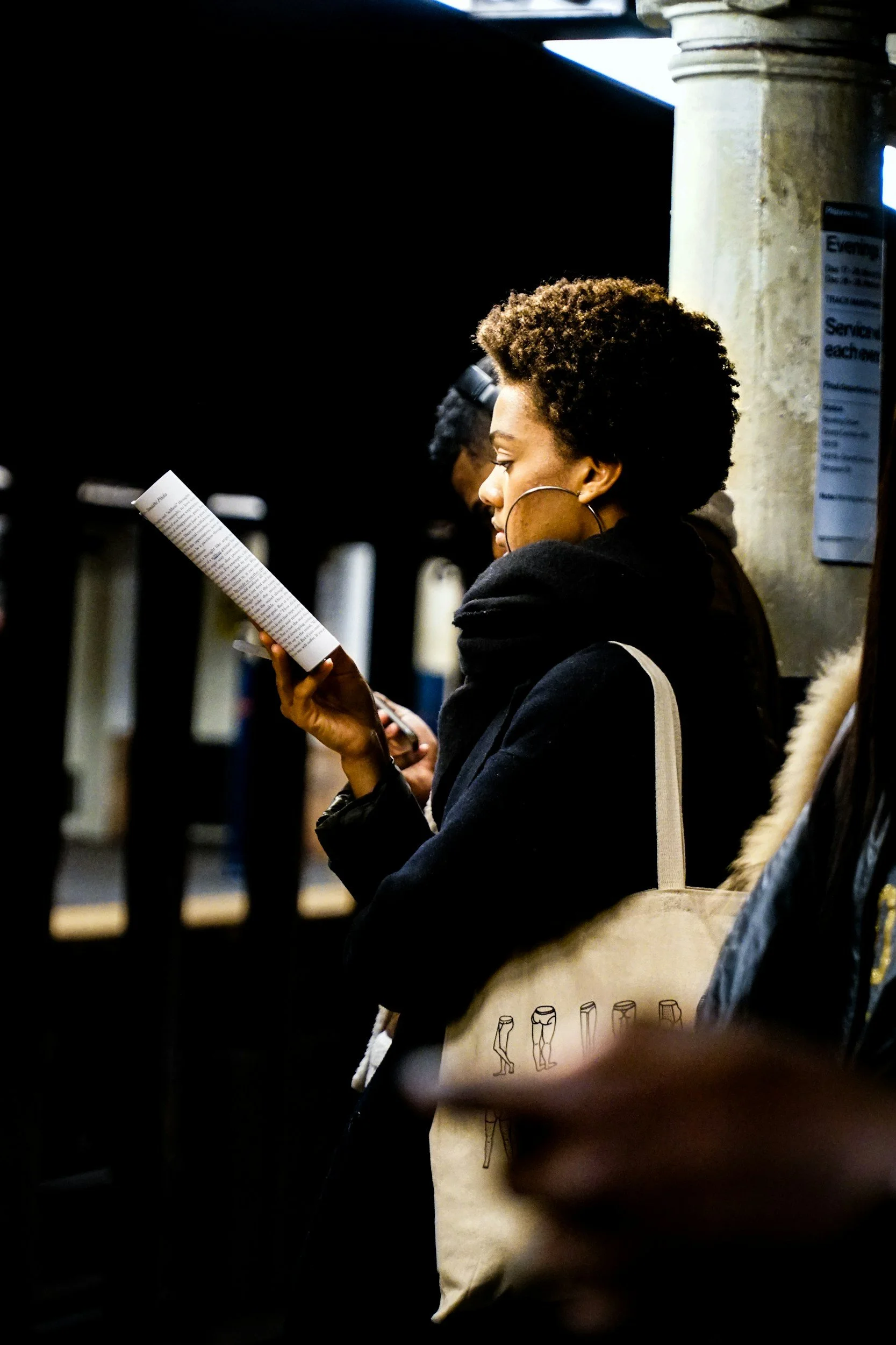 A woman waits at a subway station, reading a book while standing on a dark platform, with other commuters nearby.