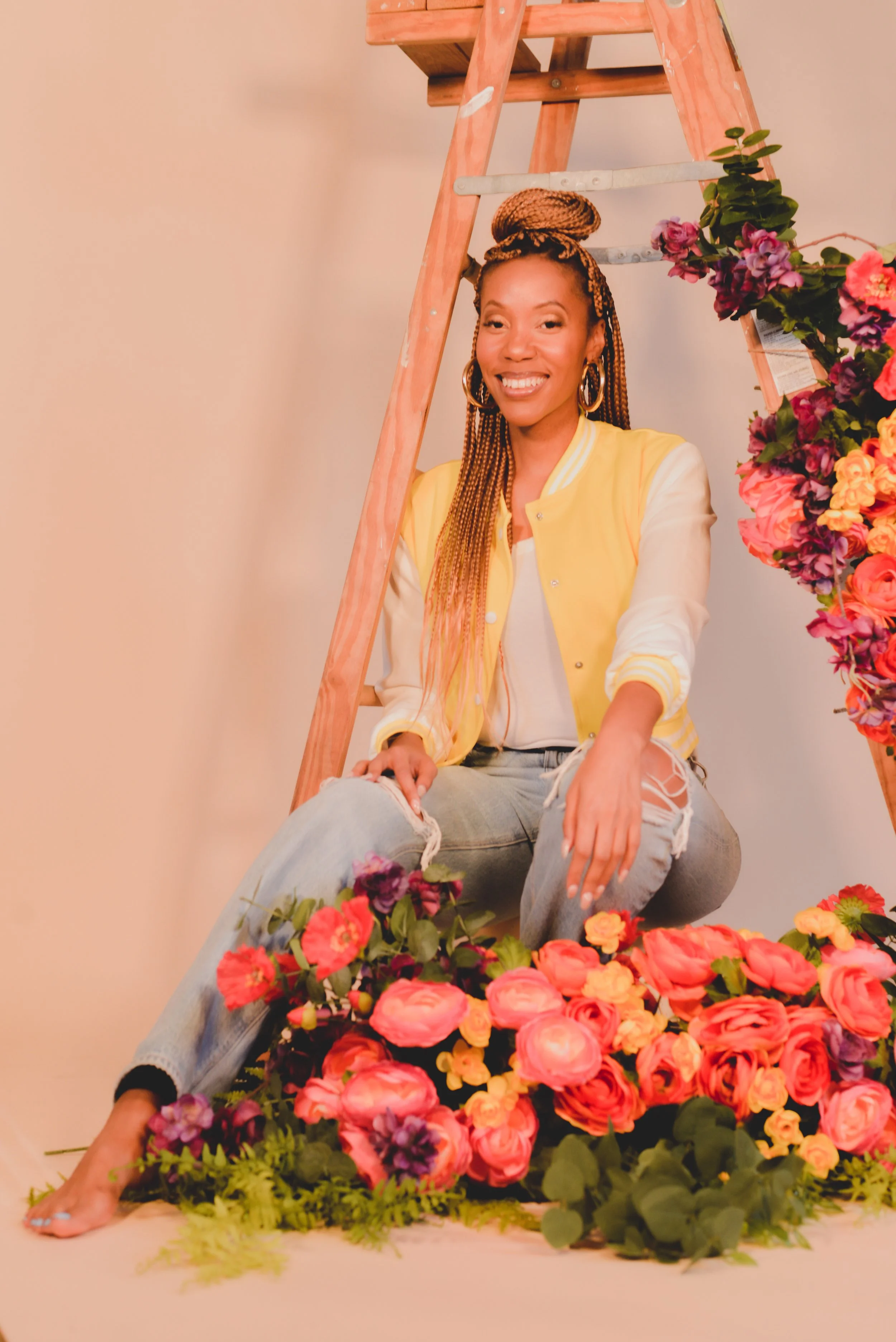 A smiling woman with long braided hair, wearing a yellow and white jacket, white shirt, and ripped jeans, sitting on the floor amidst a vibrant arrangement of pink, purple, and yellow flowers, with a wooden ladder and floral decoration in the background.