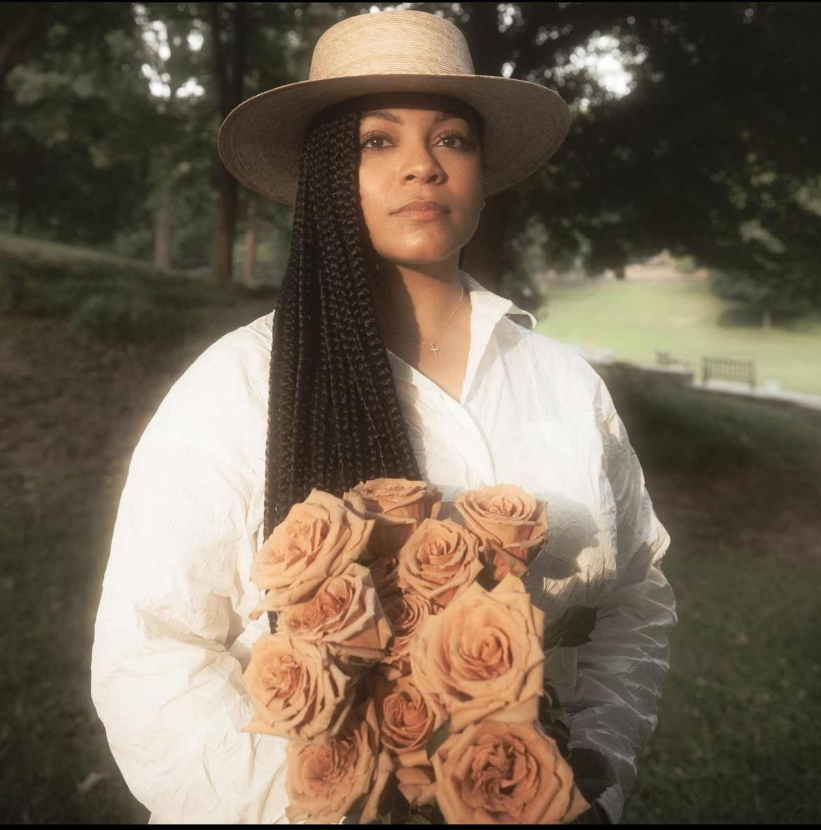 A woman with braided hair, wearing a wide-brimmed straw hat and white shirt, holding a bouquet of peach-colored roses in an outdoor park setting.