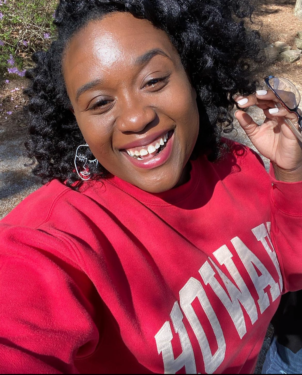 A smiling woman with dark curly hair, wearing earrings and a red sweatshirt, holds sunglasses in her hand outdoors.
