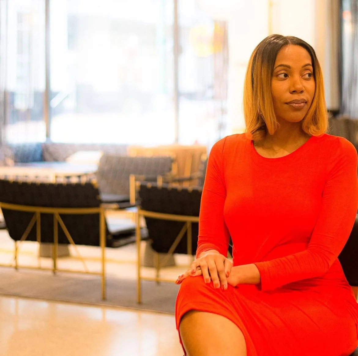 A woman with shoulder length hair wearing a red dress sitting in a well-lit, modern interior space with chairs and large windows in the background.