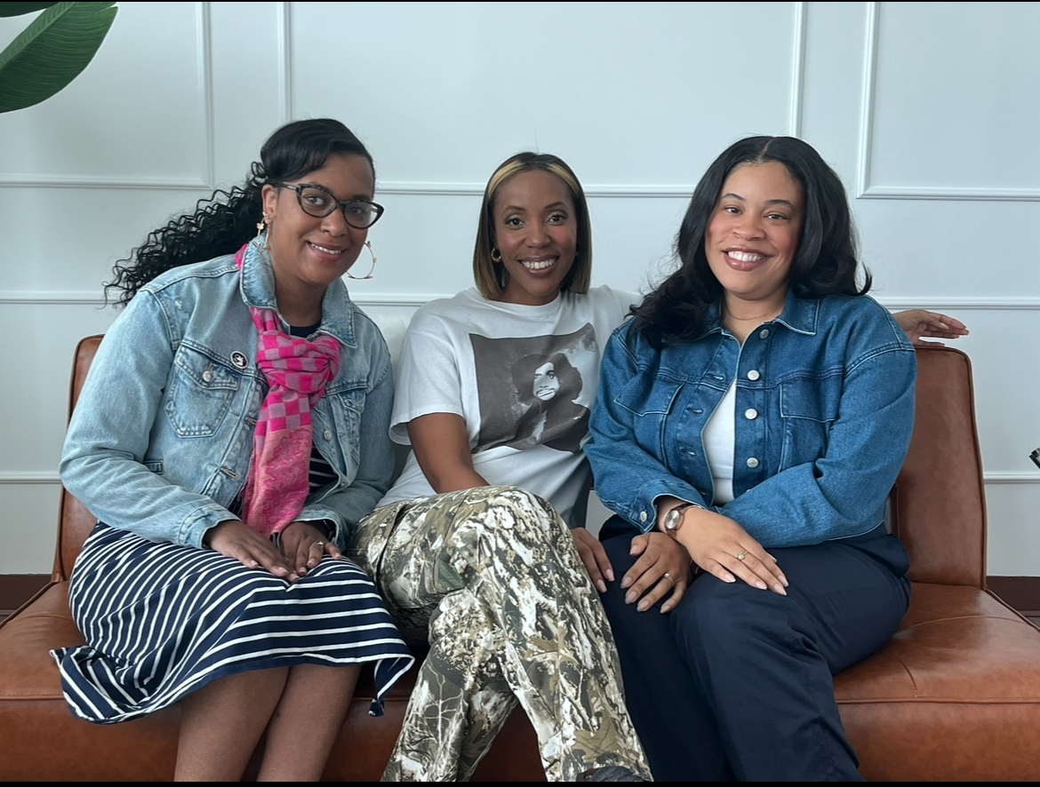 Three women sitting on a brown leather couch in a white-paneled room, smiling at the camera.