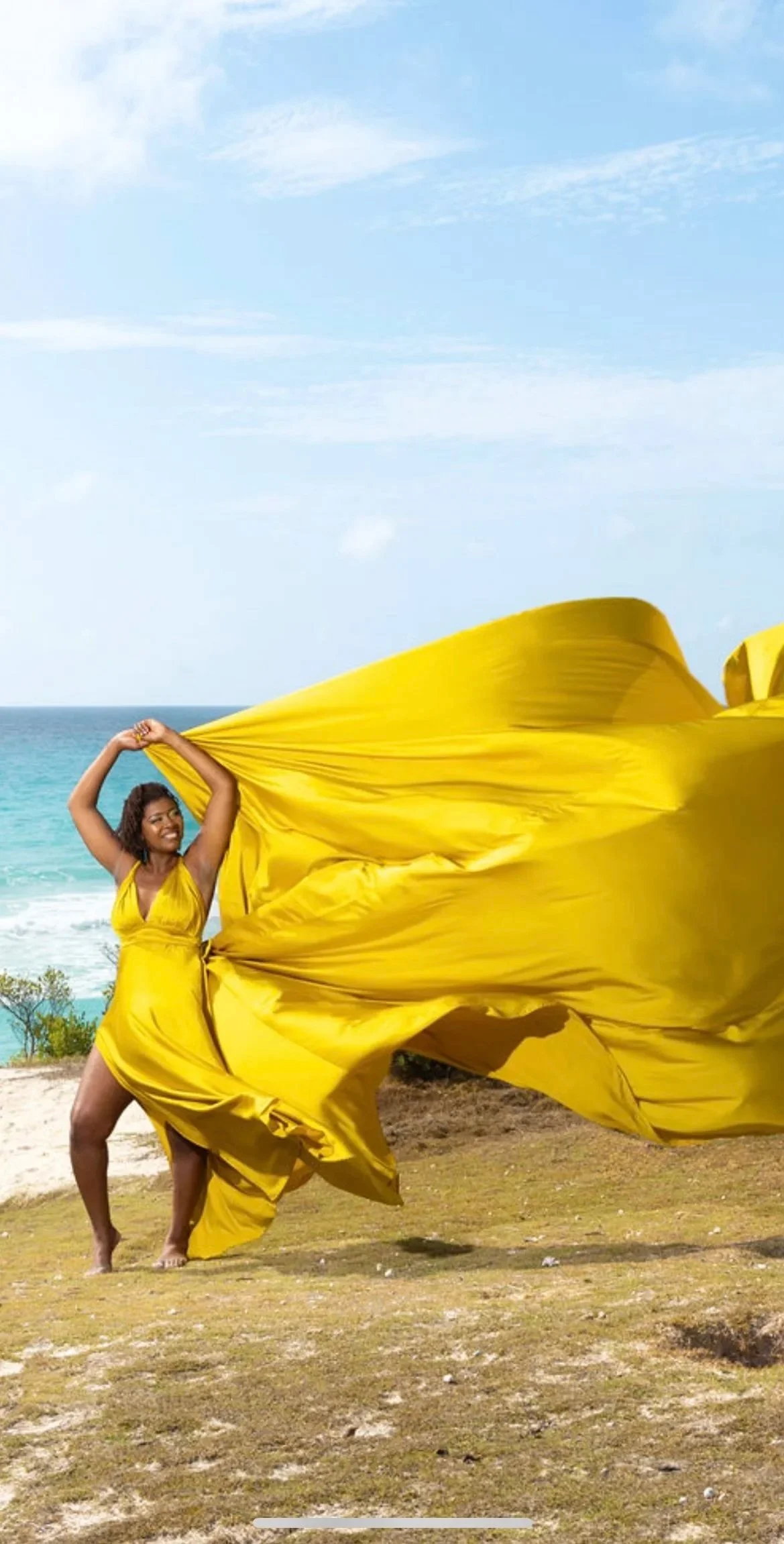 A black woman on vacation in a bright yellow flying dress stands outdoors in front of the ocean, holding and flowing the fabric of her dress in the wind.