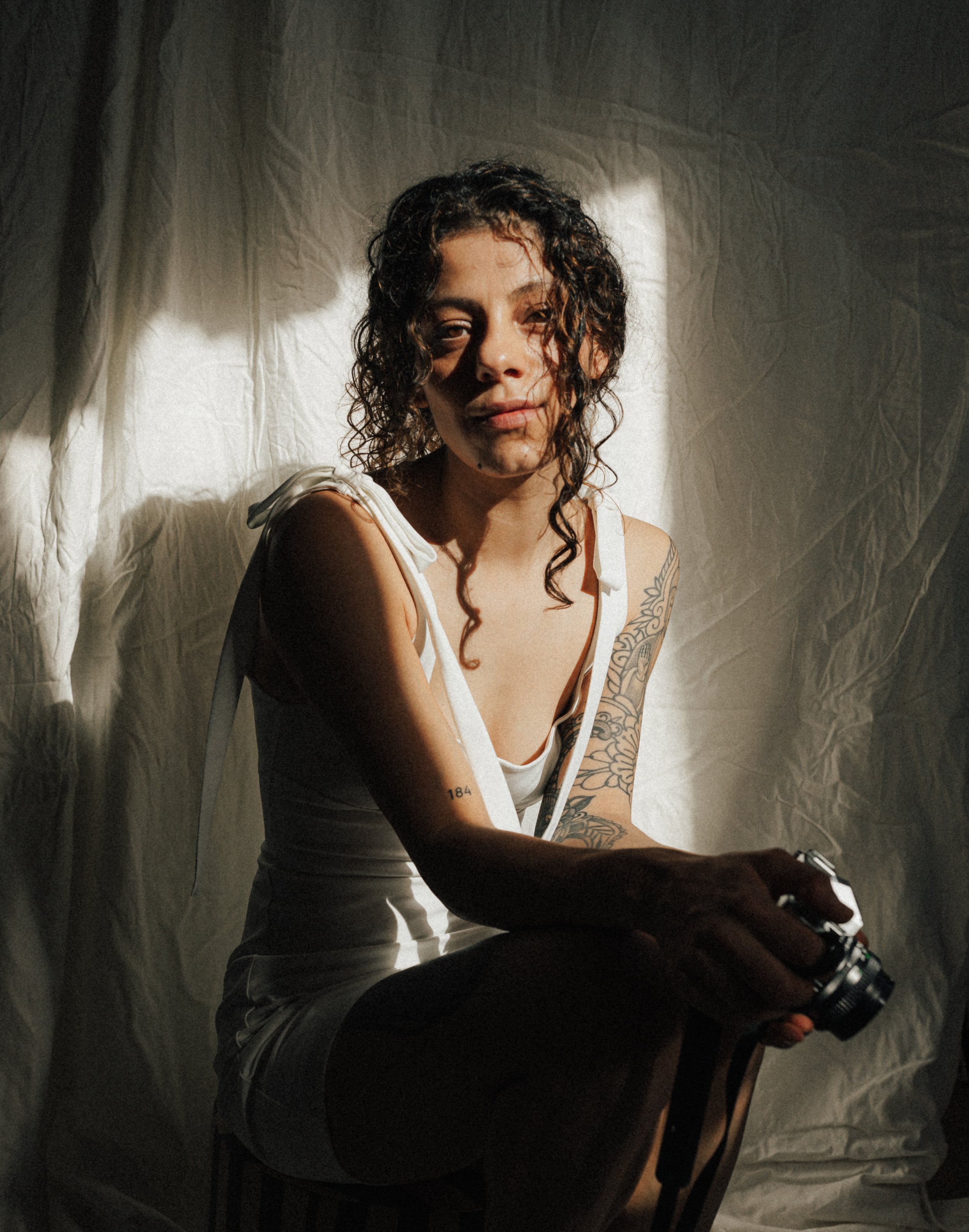 A woman with curly hair sitting in front of a white fabric backdrop, holding a camera, with sunlight casting shadows on her face.