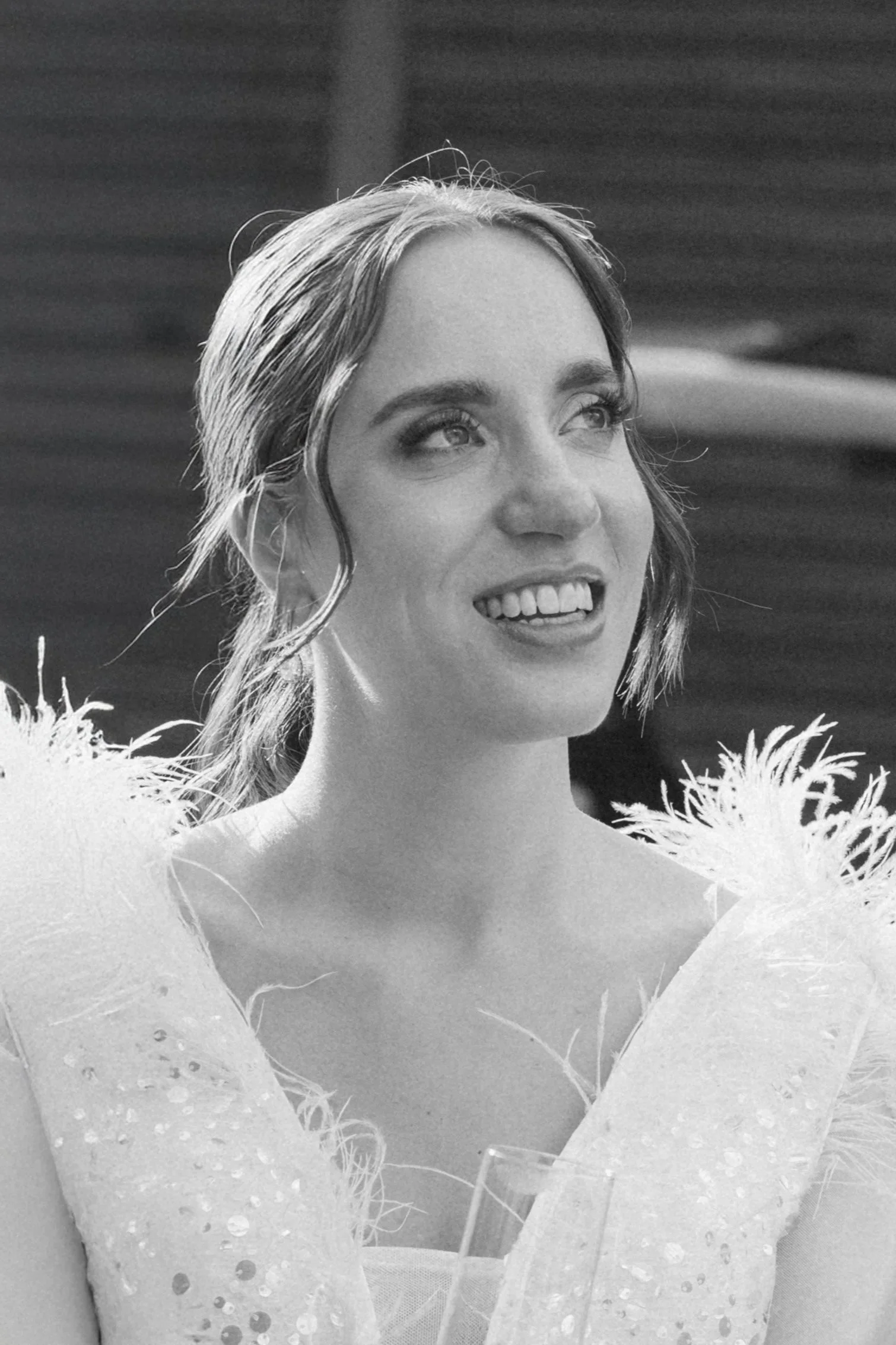 Junge Frau mit freudigem Ausdruck, lockiger Bob-Frisur, trägt ein Kleid mit Federn und Pailletten, hält ein Weinglas in der Hand, im Hintergrund eine Person und eine moderne Holzverkleidung.