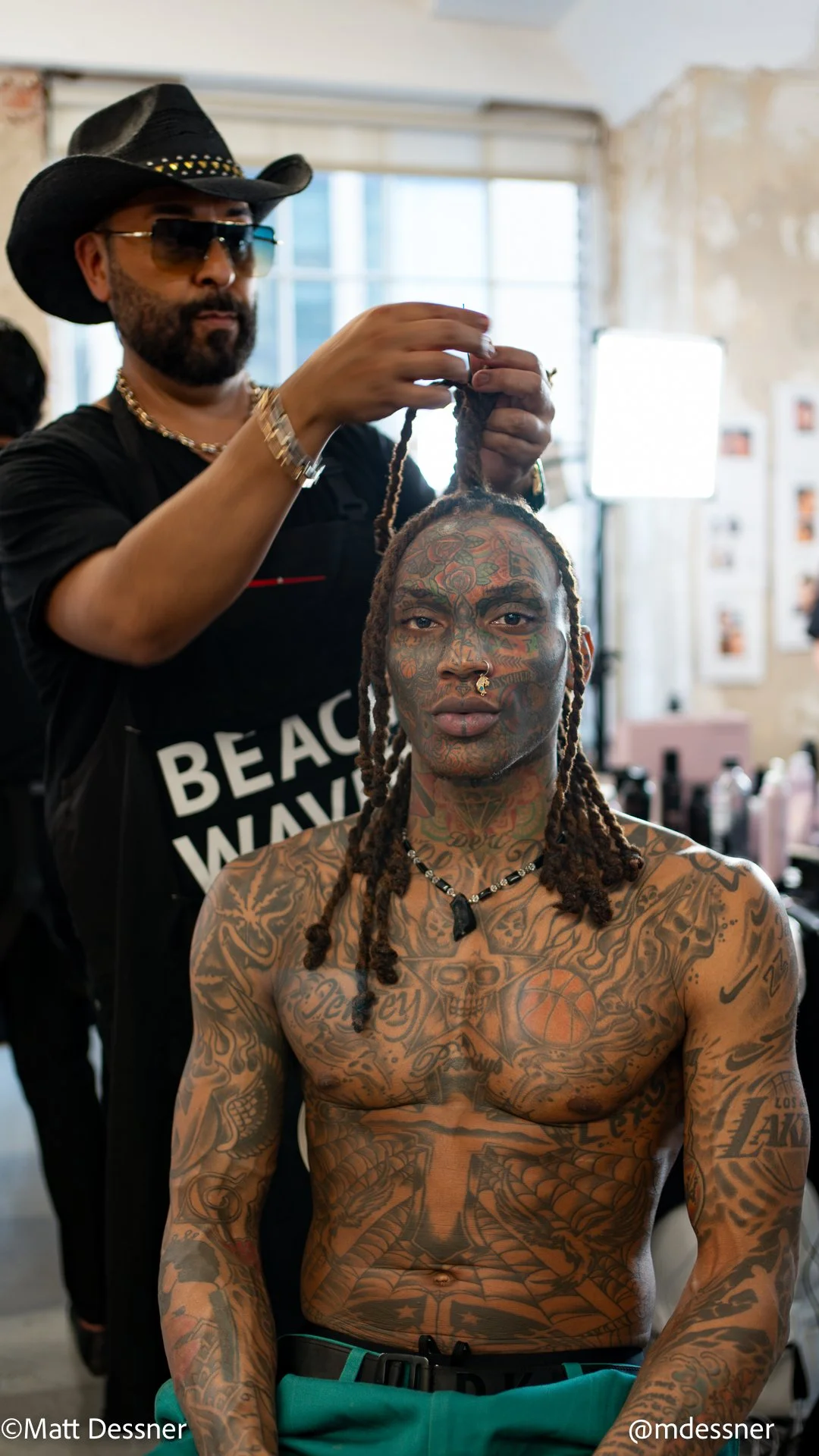 Man with tattoos, dreadlocks, and jewelry sitting shirtless while a stylist styles his hair in a studio