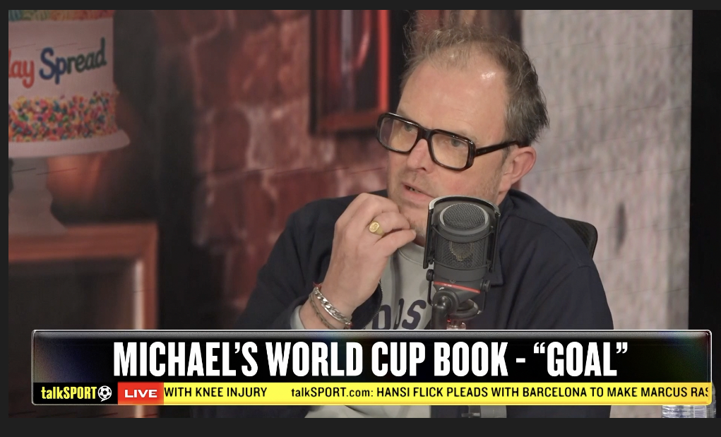 A man with glasses, a beard, and wearing a dark jacket is speaking into a microphone during a talkSPORT radio show. There is a colorful cake in the background and the caption at the bottom mentions Michael's World Cup book about the word 'goal'.