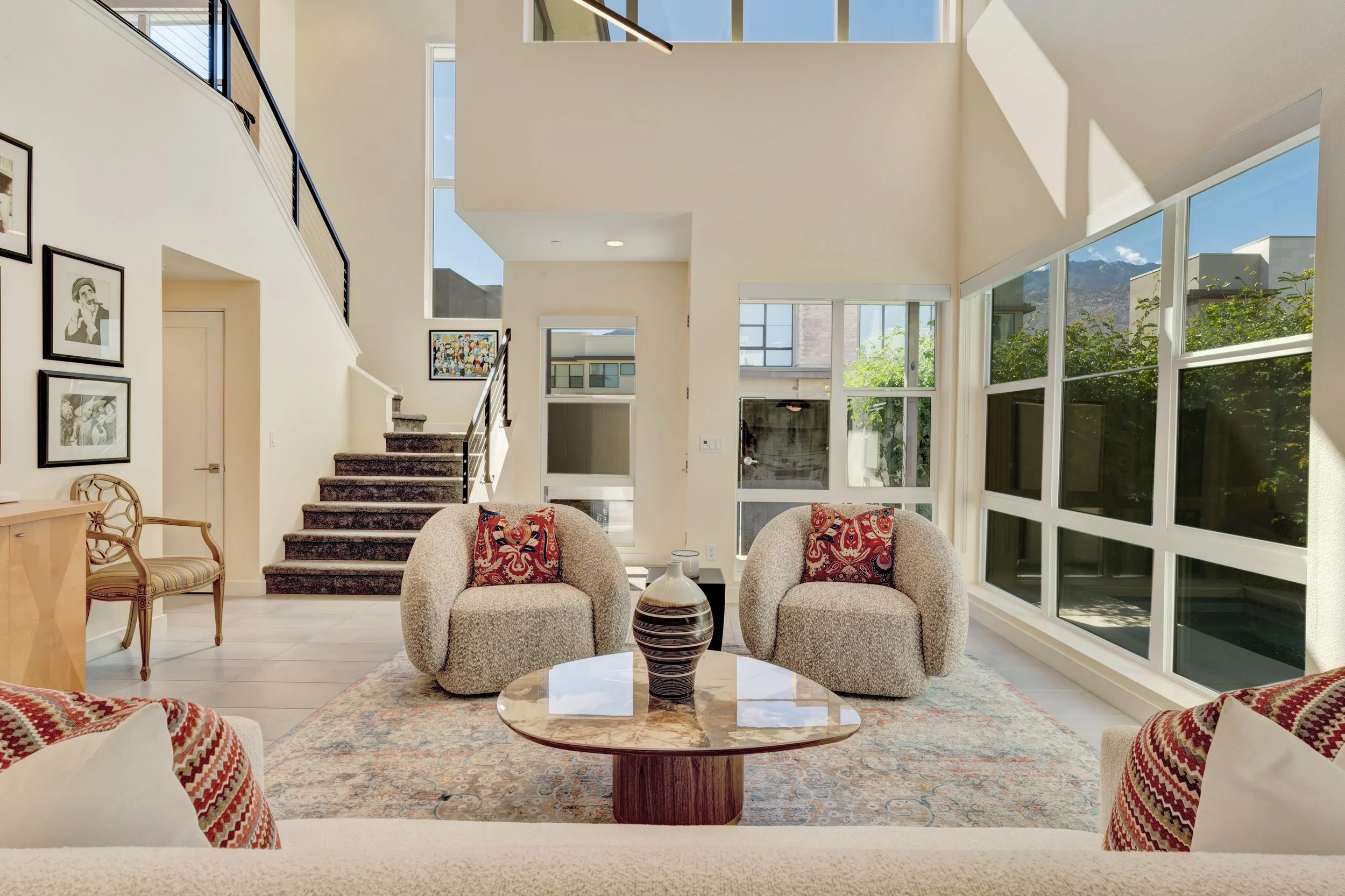 Bright living room with large windows, two beige armchairs with red patterned pillows set around a glass table with a vase, a patterned rug, and stairs leading to upper floor, decorated with black and white photographs and colorful artwork.