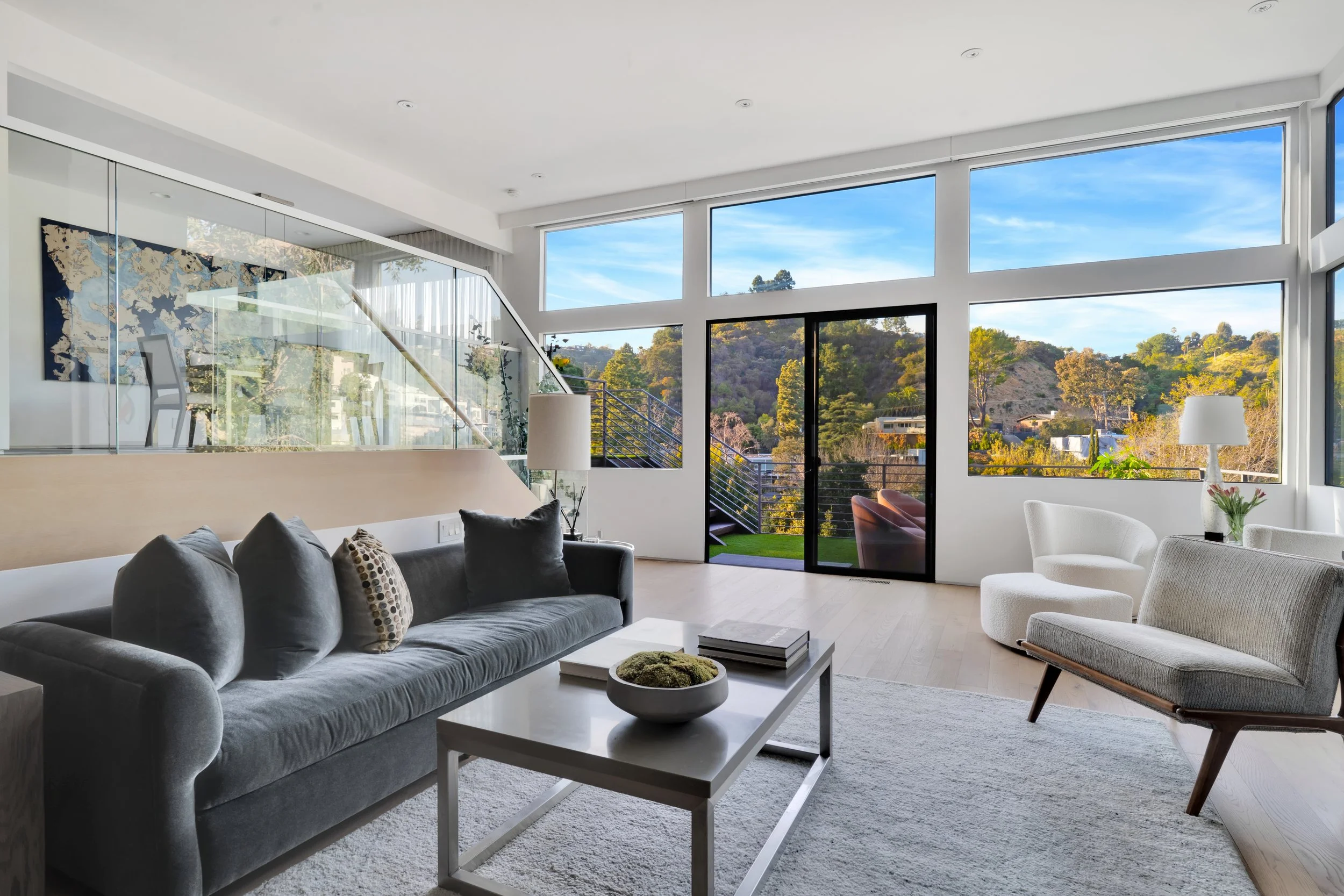 Modern living room with large windows revealing a scenic outdoor view of trees and hills, gray sofa, white armchair, coffee table with books and decorative moss, side tables with lamps, and a glass balcony door.