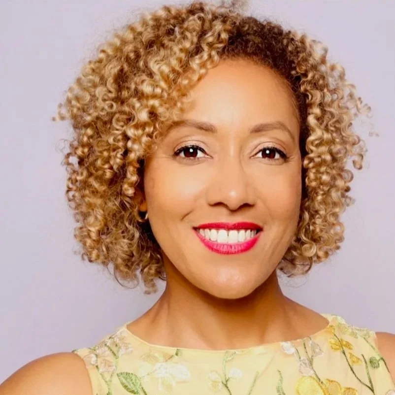 Dr. Dana Labat's professional headshot - curly blonde hair, wearing a yellow floral sleeveless top, smiling with red lipstick against a plain light background.