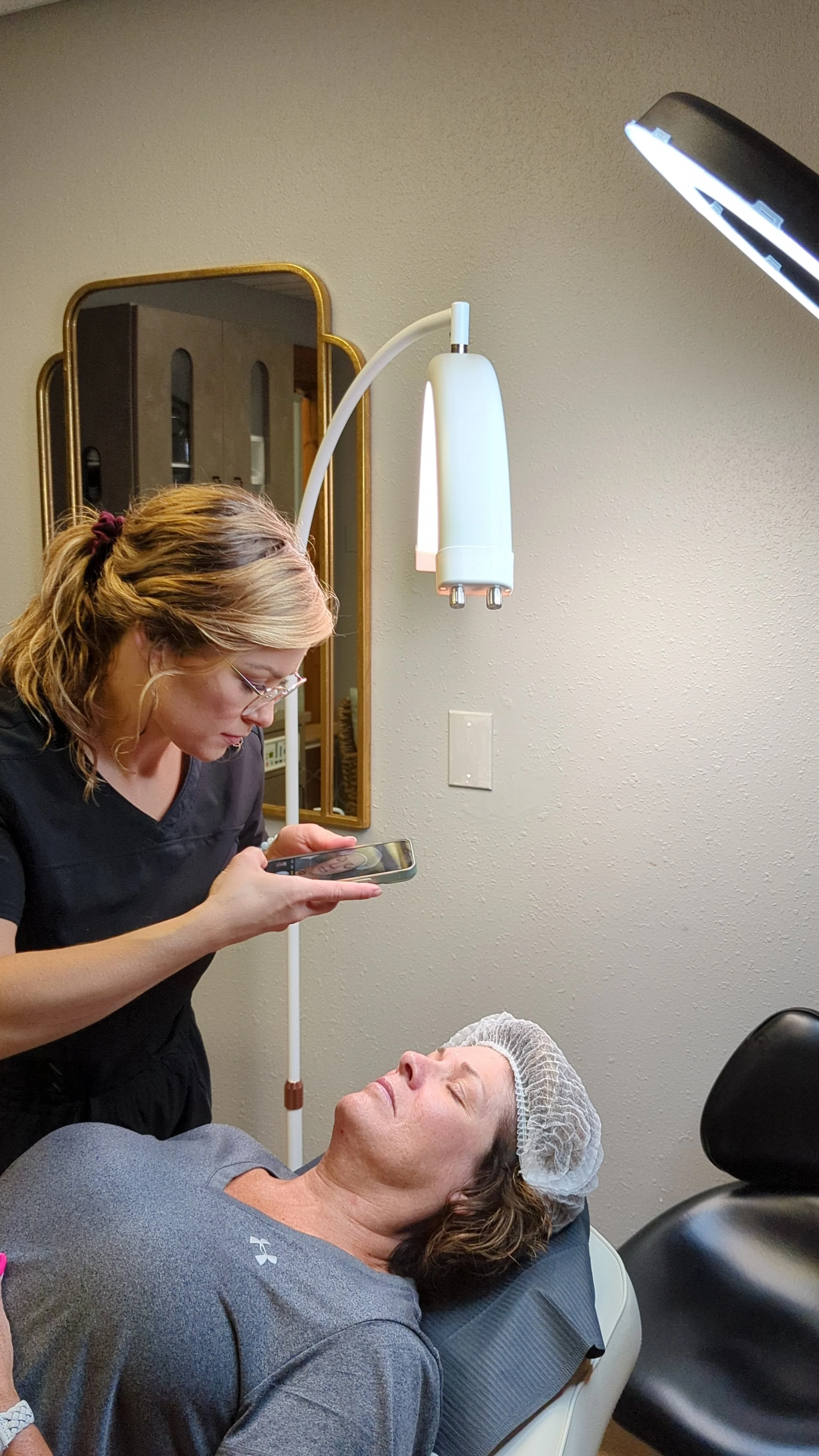 A woman in medical scrubs and glasses leans over a woman lying in a dental chair, who is wearing a hair cap and sunglasses, while the woman in scrubs looks at her phone. There is a dental light overhead and a mirror on the wall in the background.