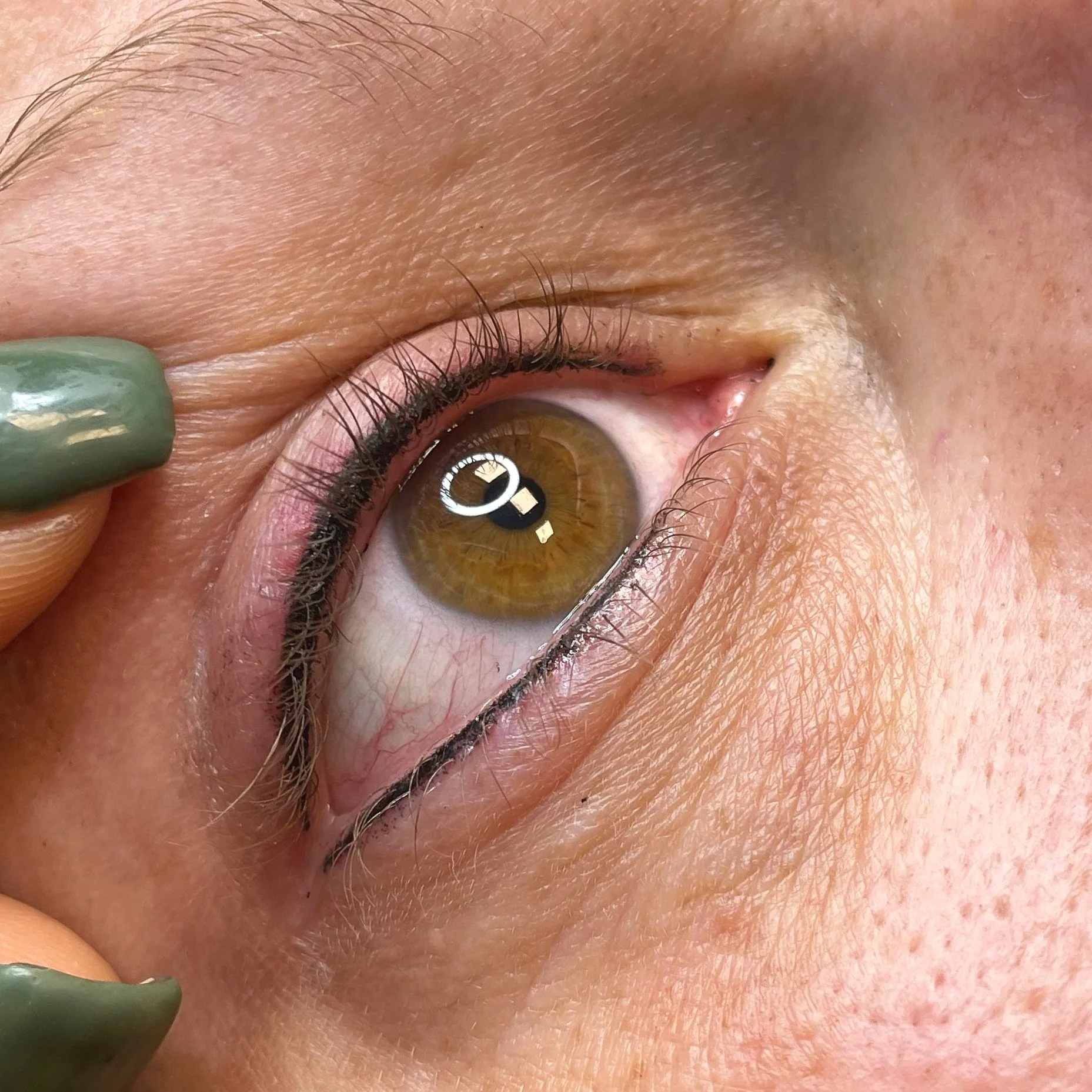 Close-up of a person's eye with brown iris, surrounded by skin, eyelashes, and a hint of eyebrow. A hand with painted green nails is gently pulling down the lower eyelid.