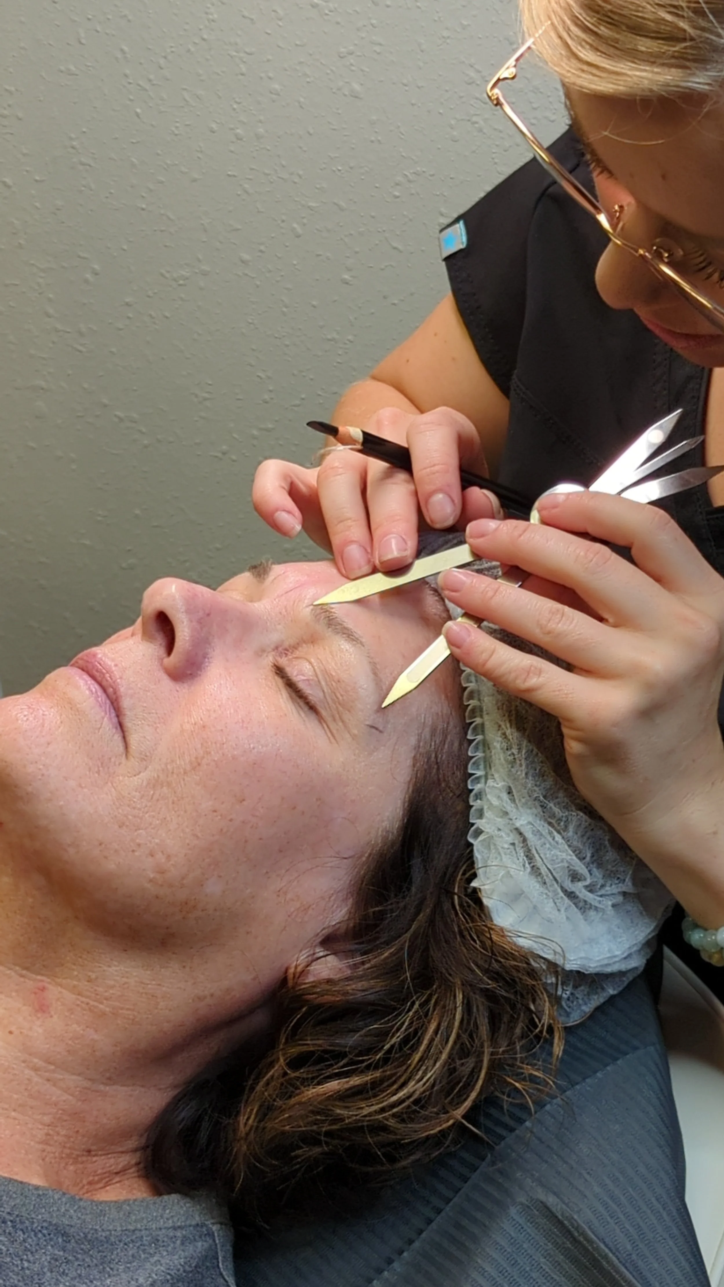 A woman receiving a cosmetic procedure on her eyebrows from a professional using small tools.