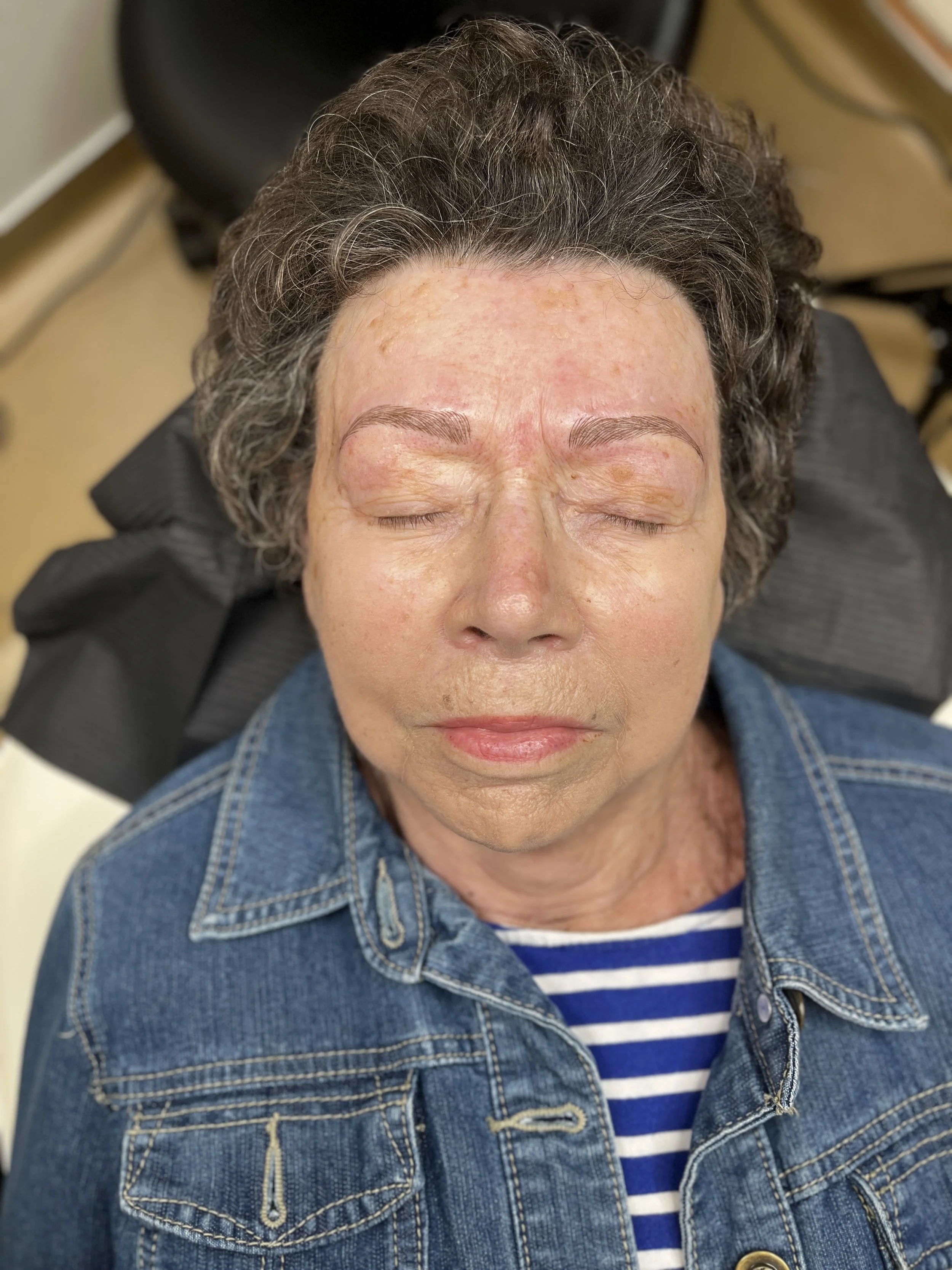 A woman with short curly gray hair and closed eyes, wearing a denim jacket over a striped shirt, lying back with her head supported on a chair or recliner.