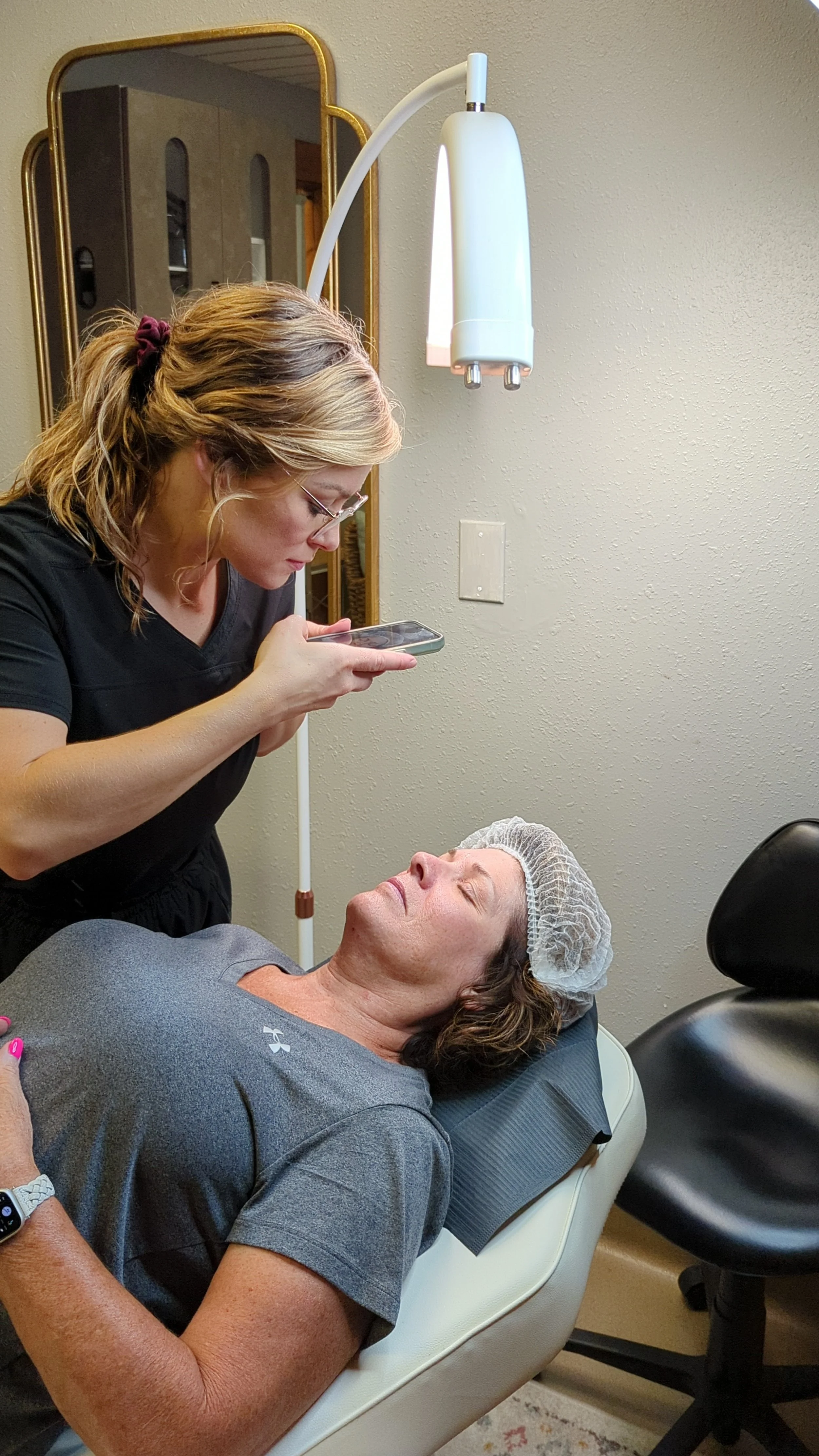 A woman lying on a dental chair with a hair cap, receiving a facial treatment from a healthcare professional. The professional is looking at her phone while examining her face.