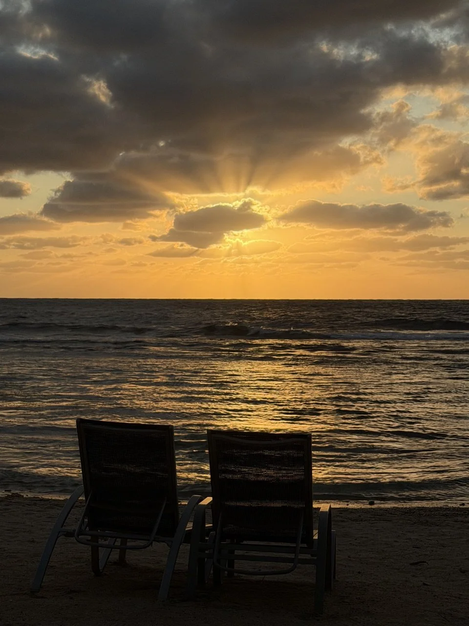 Sunset over the ocean with two empty chairs on the beach, partly cloudy sky.