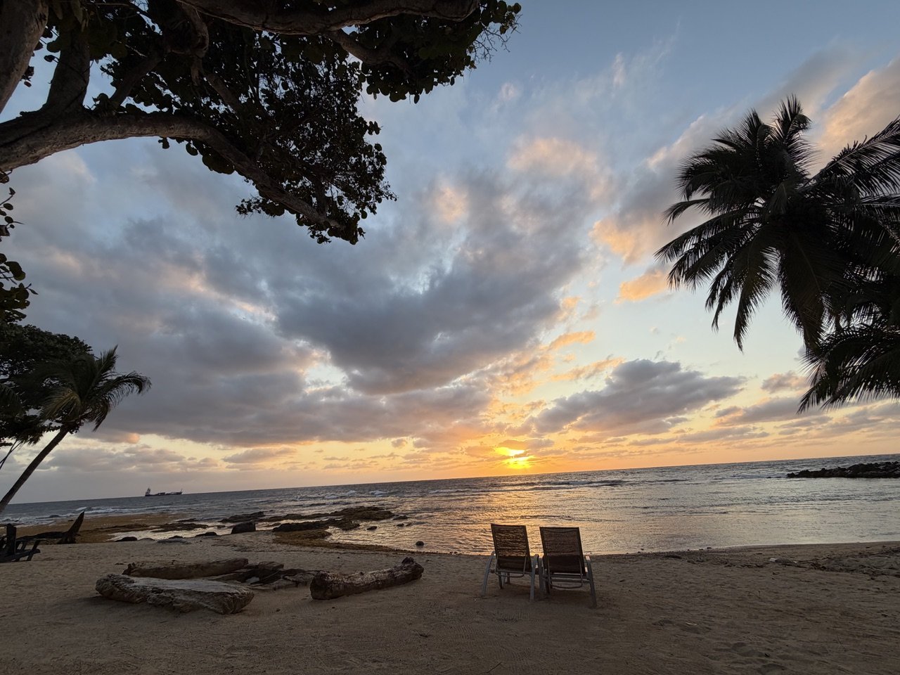 Sunset over the ocean with two chairs on the sandy beach, framed by palm trees, partly cloudy sky.