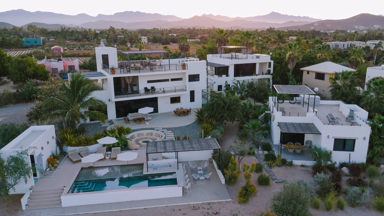 Aerial view of a modern white residential complex with multiple balconies and terraces, a swimming pool with umbrellas, lush greenery, and desert landscape with mountains in the background.