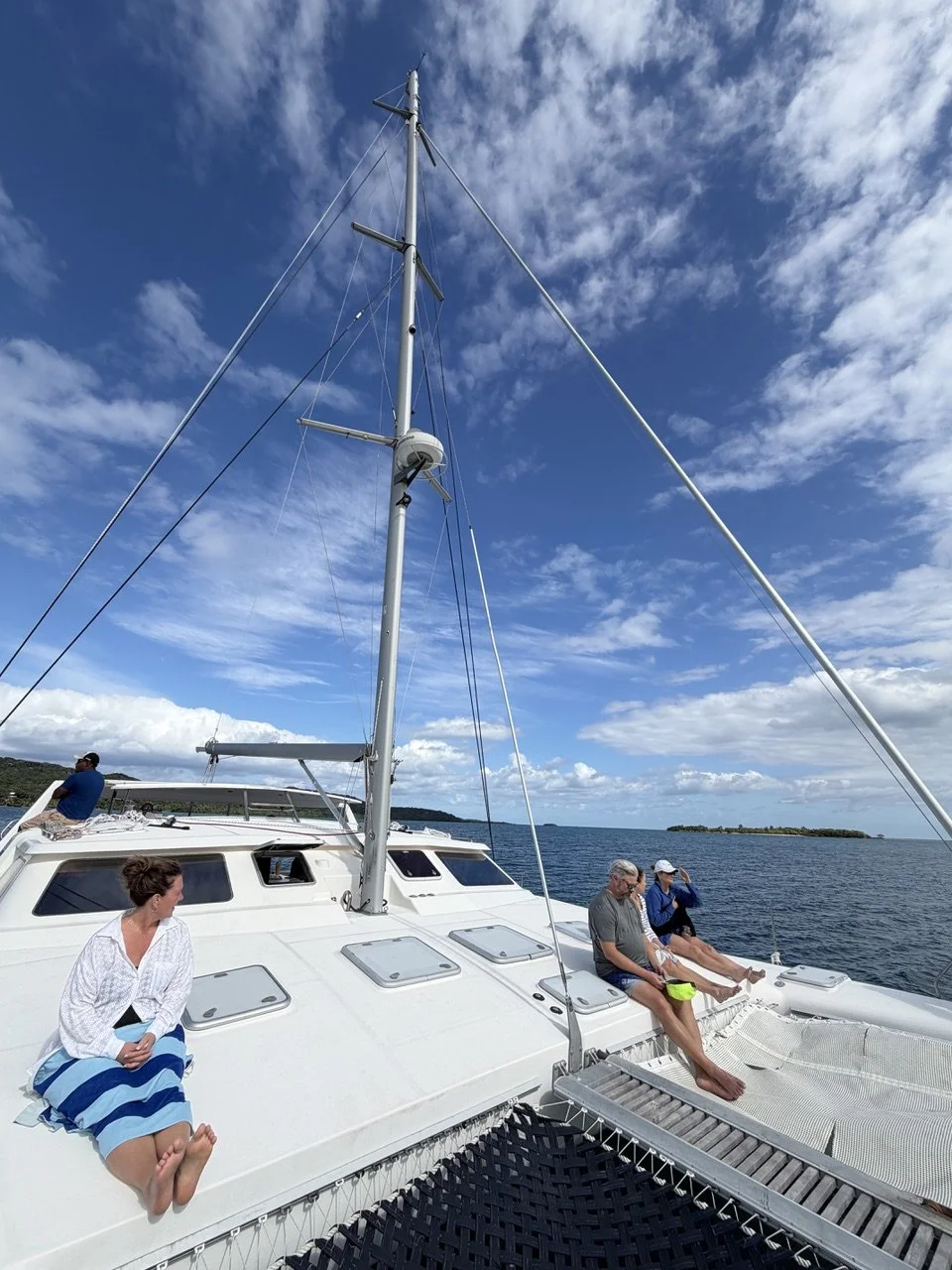 Four people relaxing on a white sailboat with a blue sky and calm waters, with small islands in the distance.