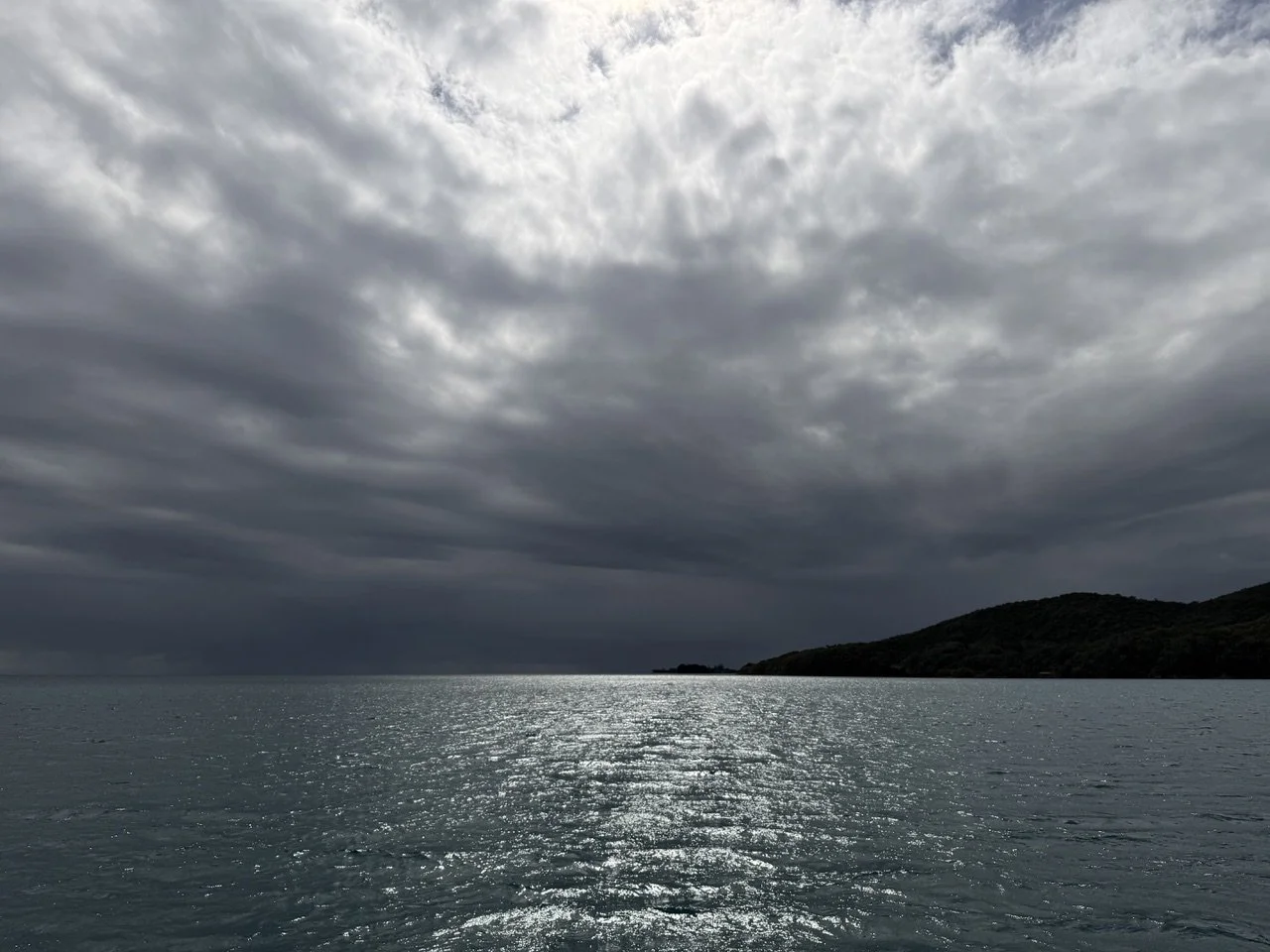 Overcast sky with dark clouds reflecting on a body of water, distant shoreline with hills.