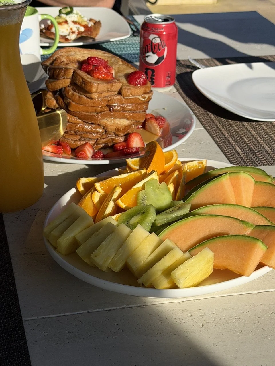 A white plate filled with cutmelon slices, kiwi slices, orange segments, and pineapple pieces on an outdoor table. In the background, there is a plate with French toast topped with strawberries, a can of Coca-Cola, and a pitcher of juice.