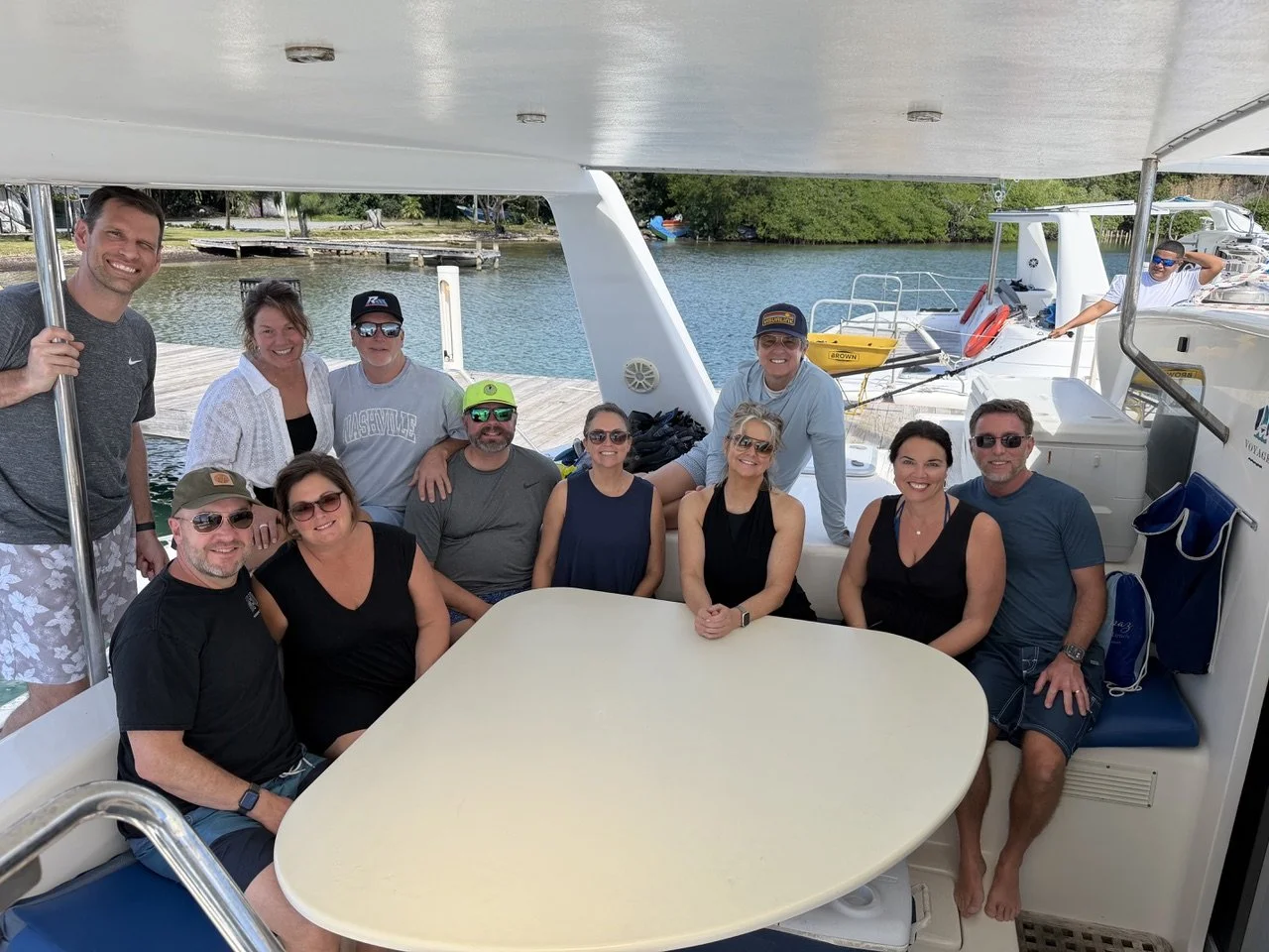 Group of twelve people on a boat docked near water, smiling for the camera, with trees and boats in the background.