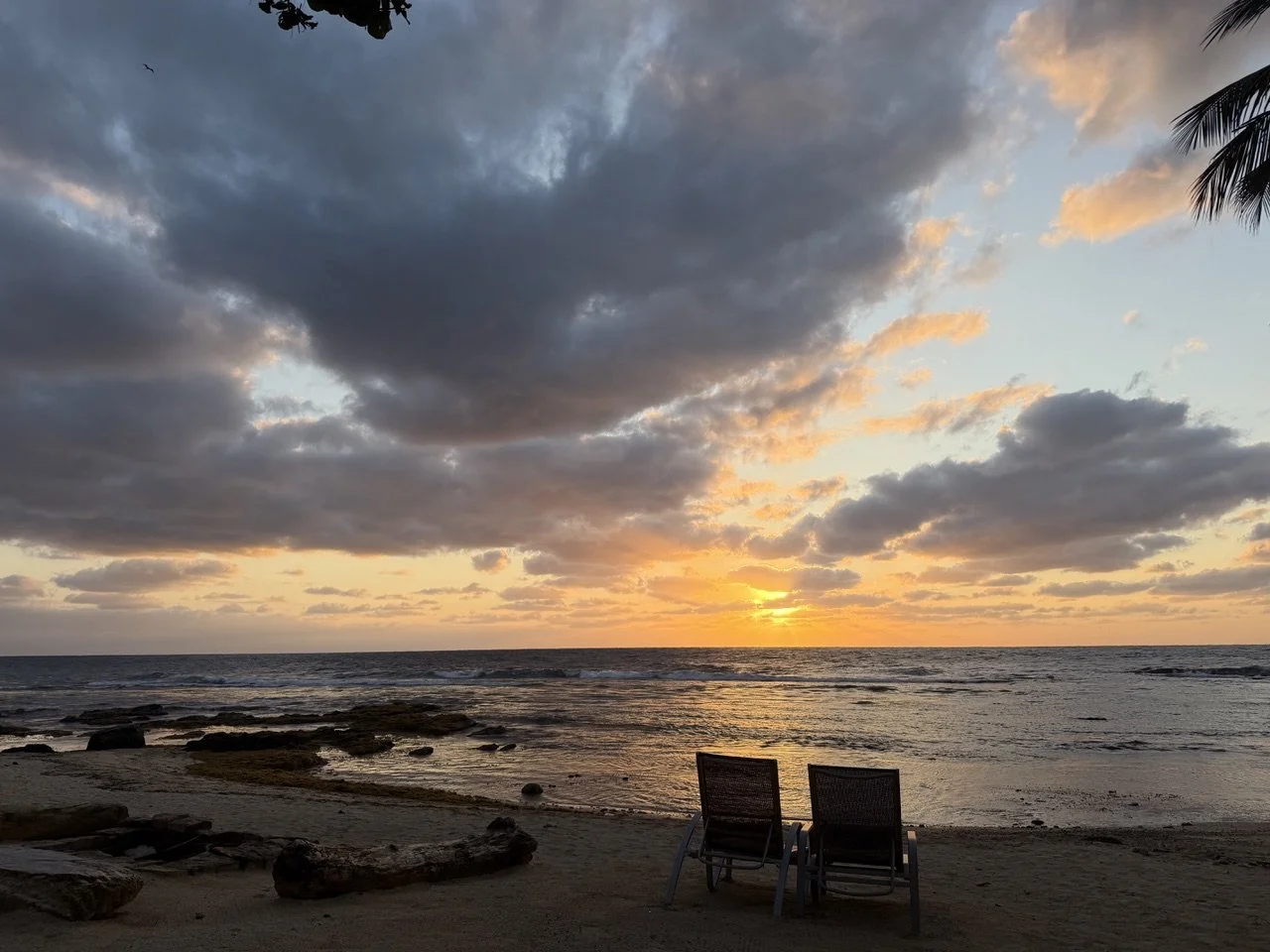 Two lounge chairs on a sandy beach at sunset, with a cloudy sky and palm leaves visible at the top right corner.