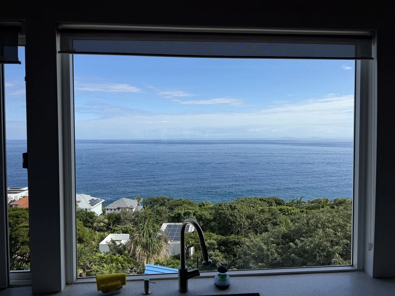 View of the ocean through a window with houses and greenery in the foreground.