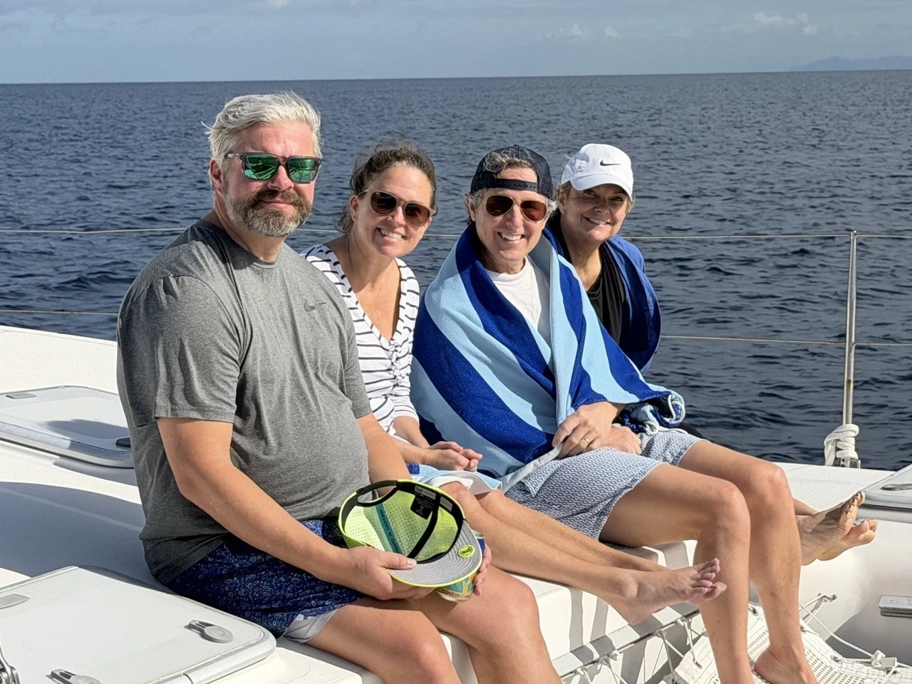 Four people sitting on a boat in open water, smiling at the camera, with two women and two men wearing casual summer clothing and sunglasses, some with towels wrapped around their shoulders.