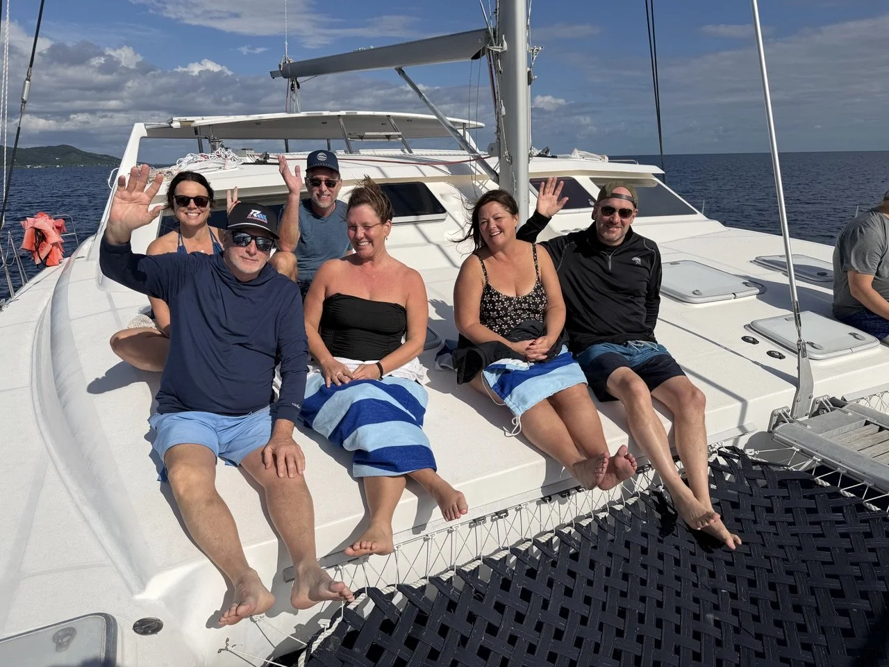 Group of six people sitting on the deck of a yacht, smiling and waving, with ocean and sky in the background.