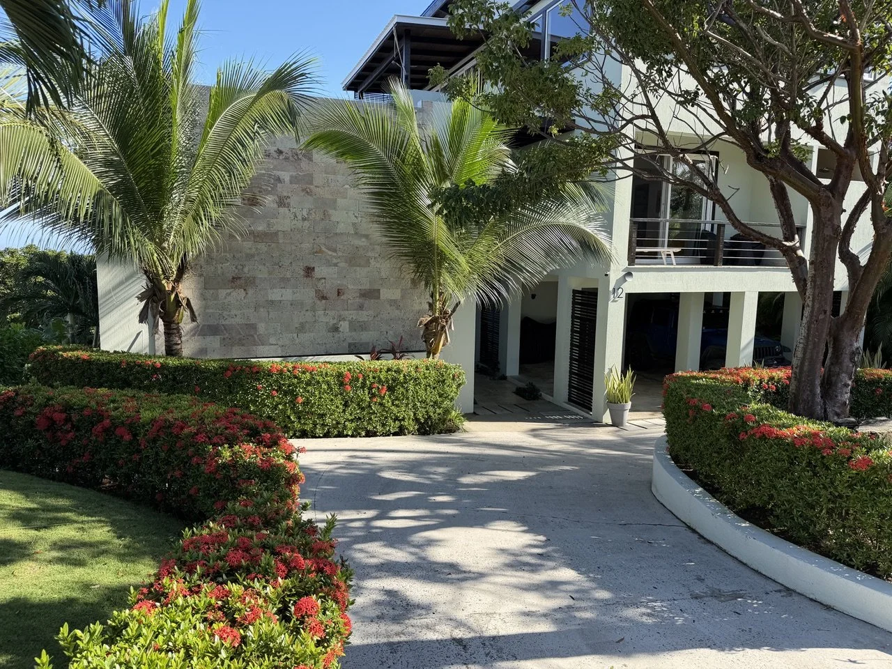 A modern house with tropical landscaping, including palm trees, colorful flowering bushes, and a well-maintained lawn. The house has a parking area beneath a balcony, with a driveway leading up to it.