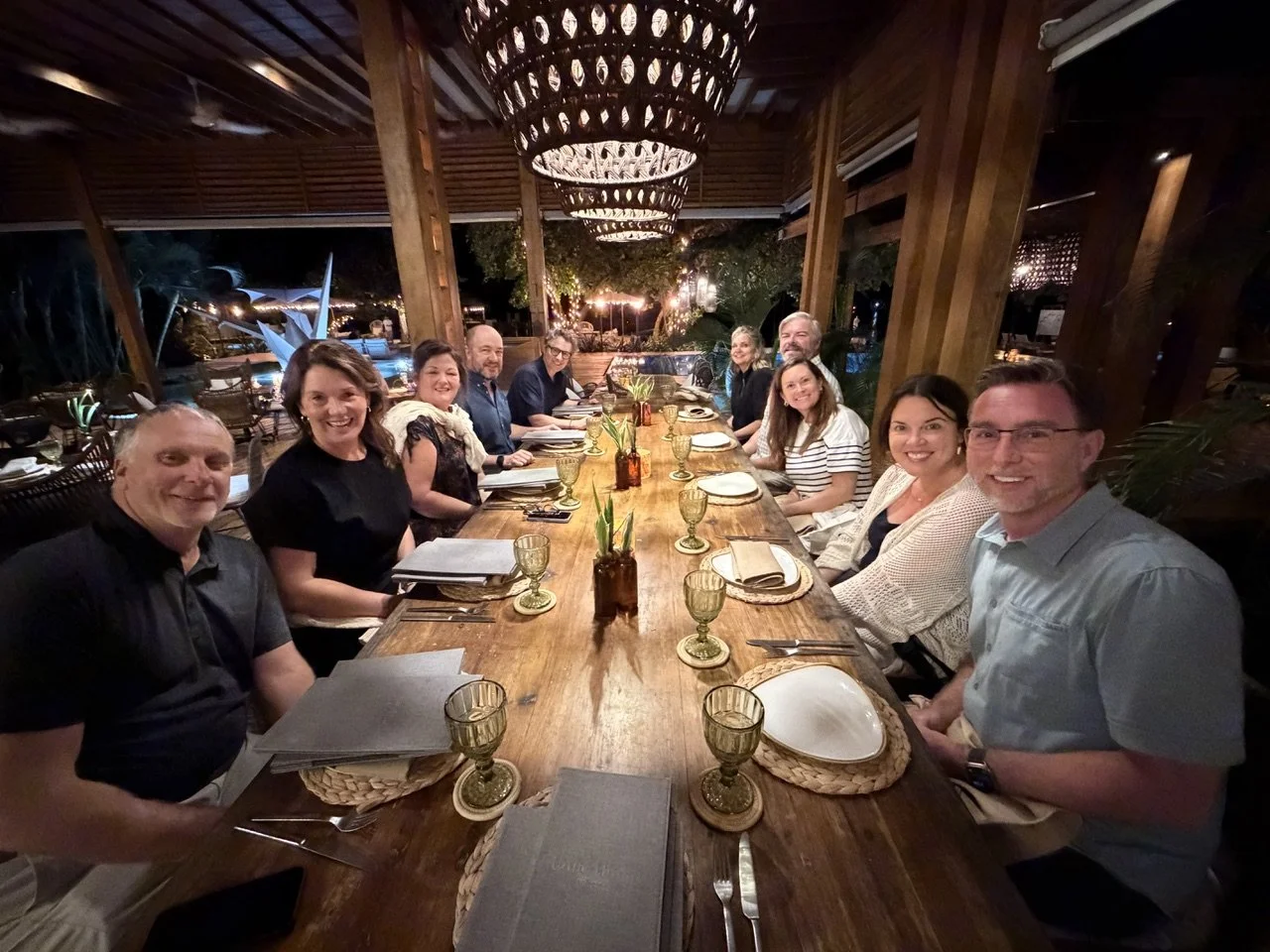 A group of ten people sitting at a long wooden dining table in an open-air restaurant at night, with a chandelier hanging above and outdoor scenery visible in the background.