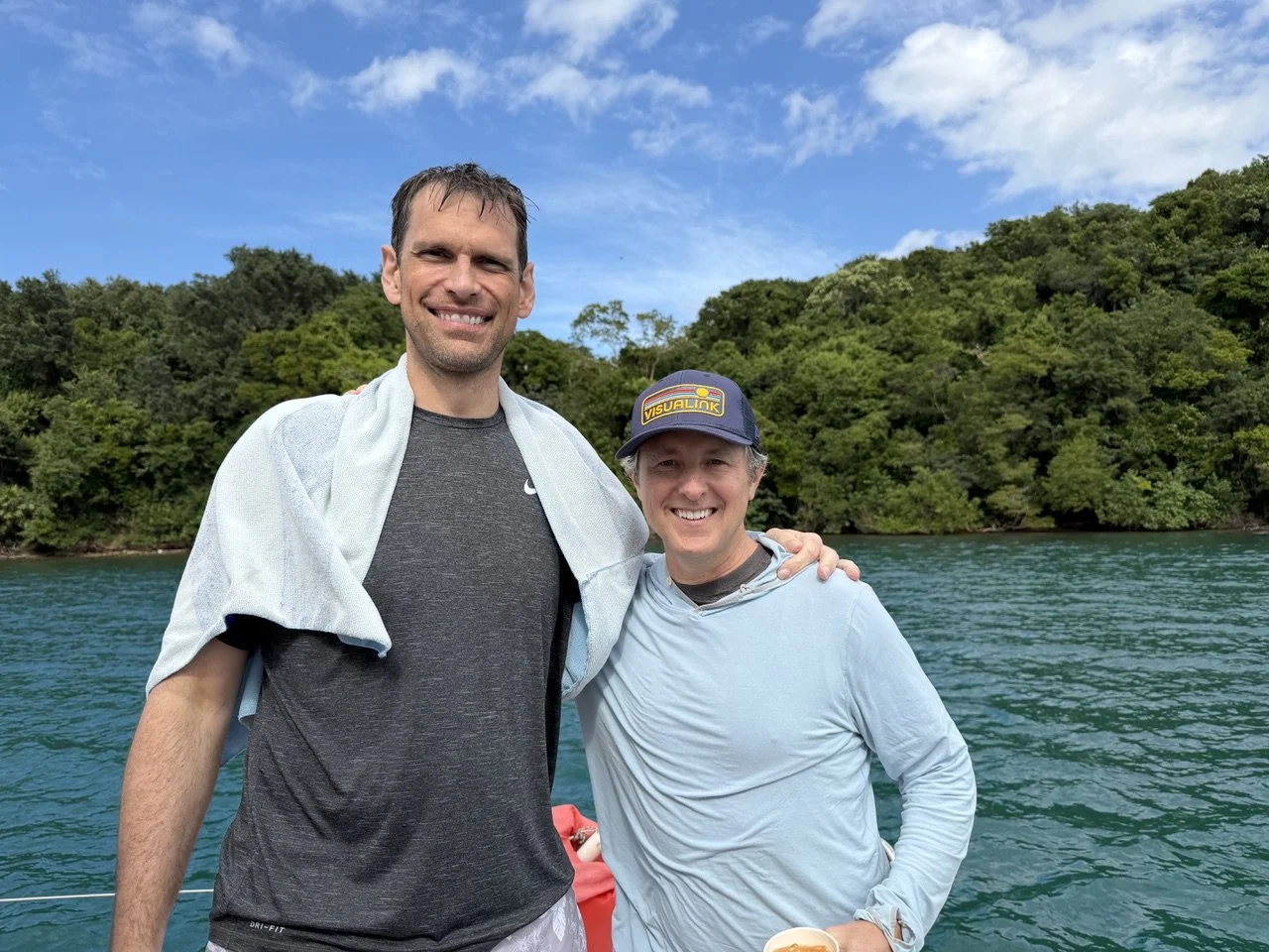 Two men standing on a boat near a lake with green trees and a partly cloudy sky in the background, smiling and posing for the photo.
