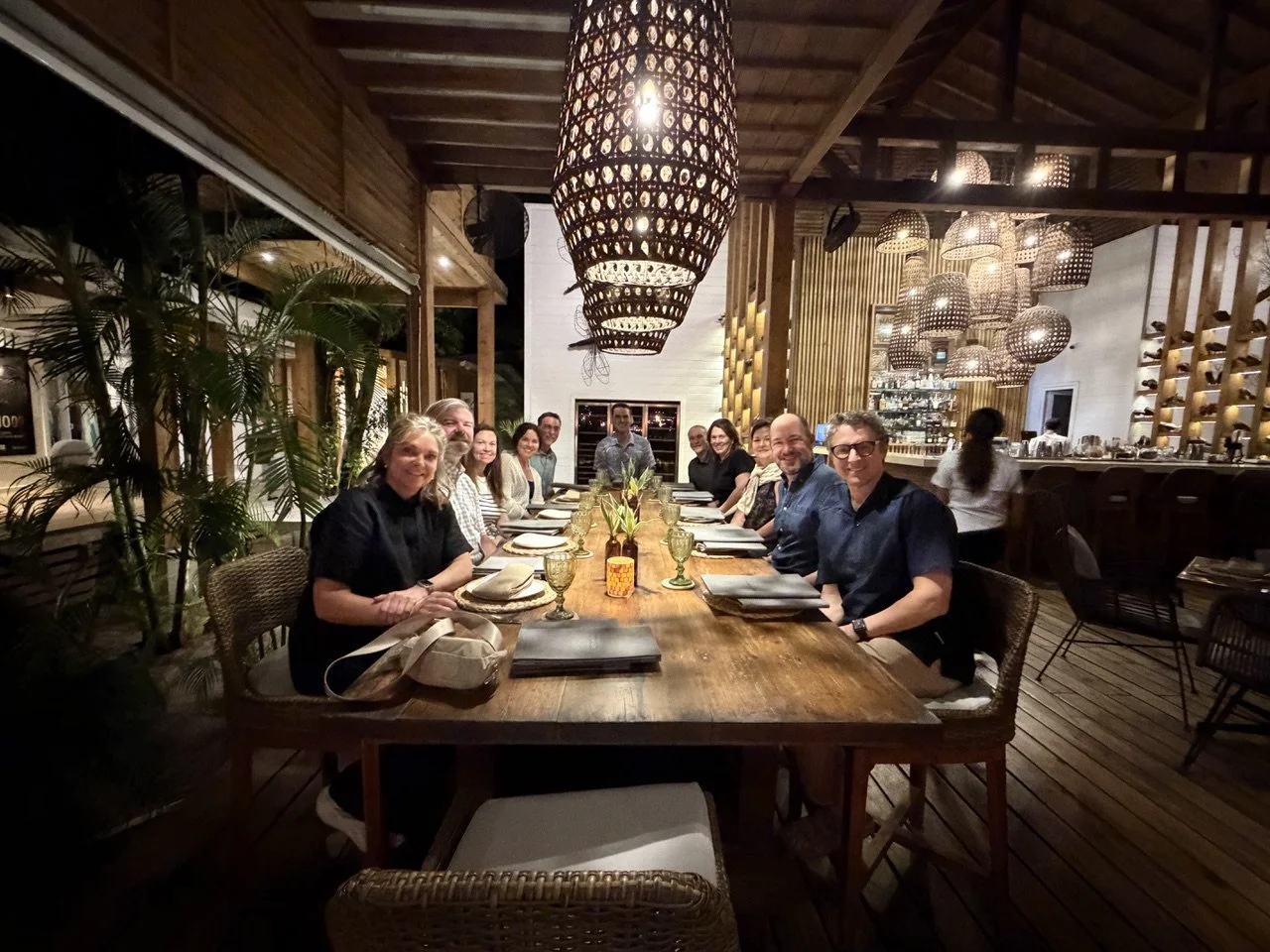 Group of people sitting at a long dinner table in a restaurant with dim lighting and wooden decor, including hanging woven light fixtures and indoor plants.