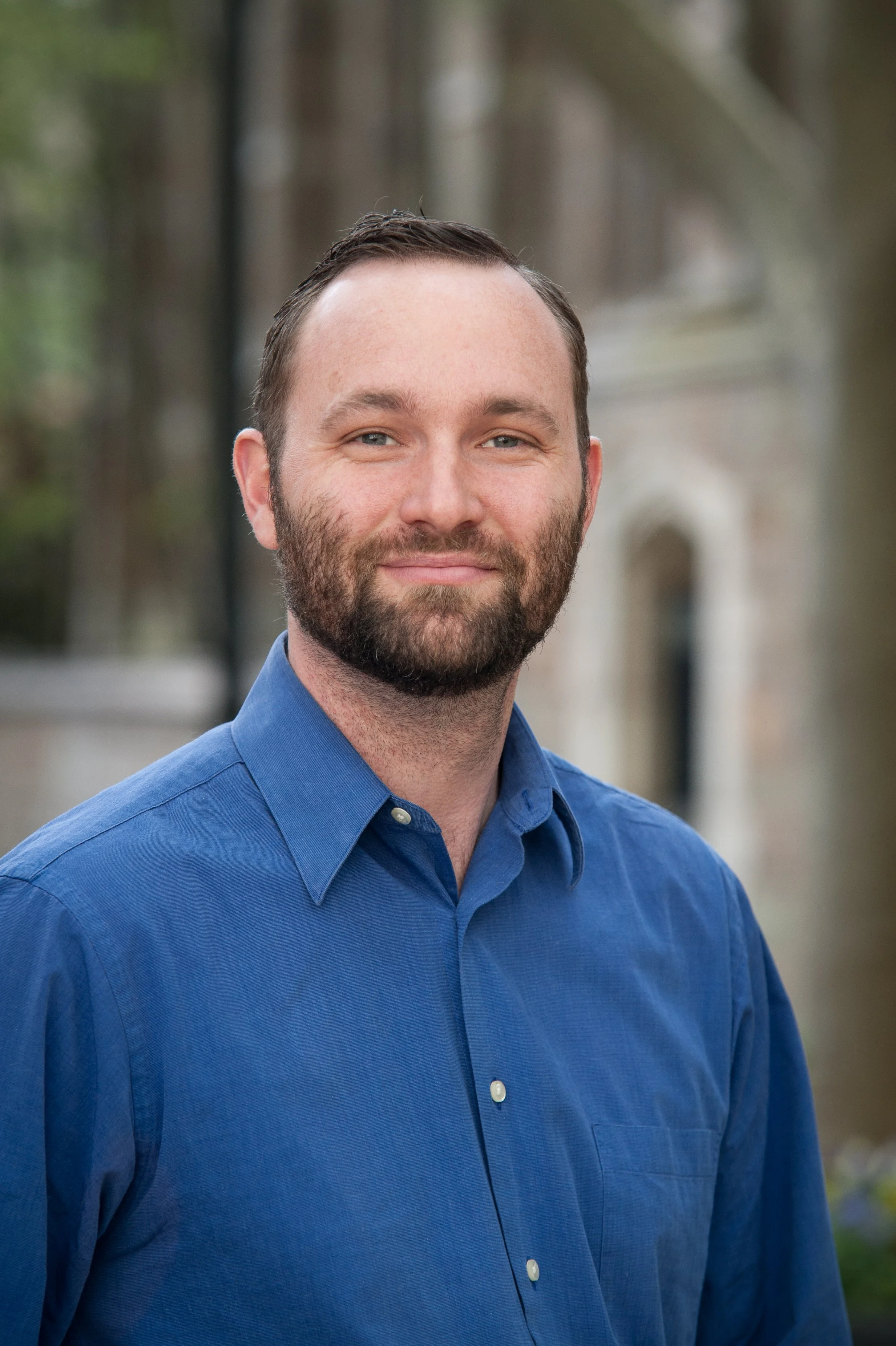 A man with a beard smiling outdoors wearing a blue buttondown collared shirt