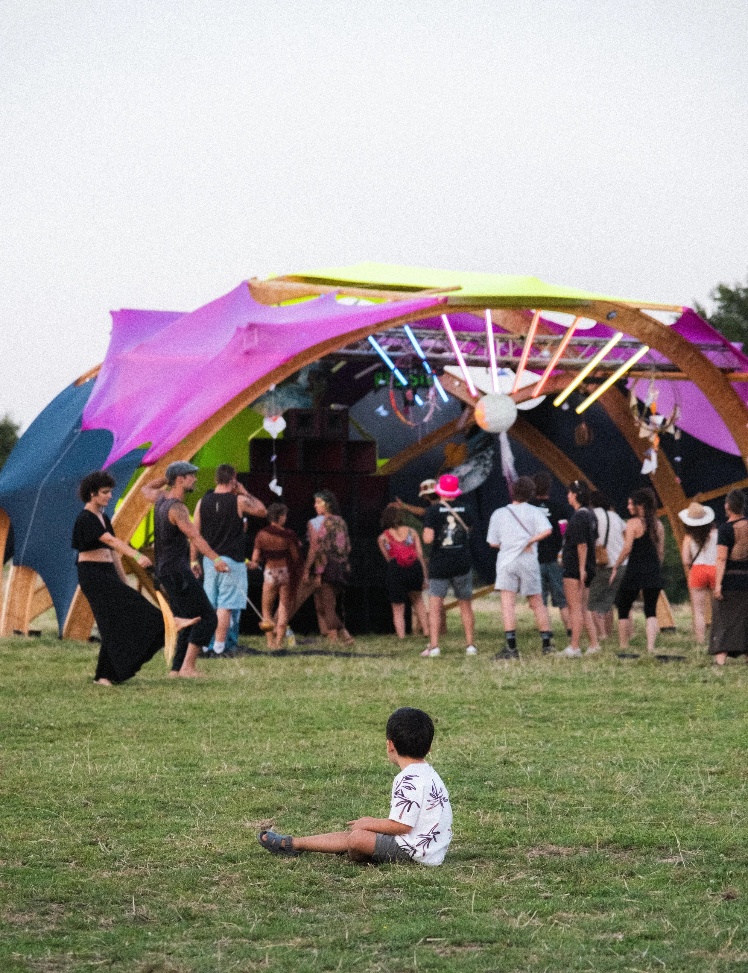 Enfants et adultes réunis sous une structure colorée lors d'un événement en plein air, avec une scène en arrière-plan, en soirée.