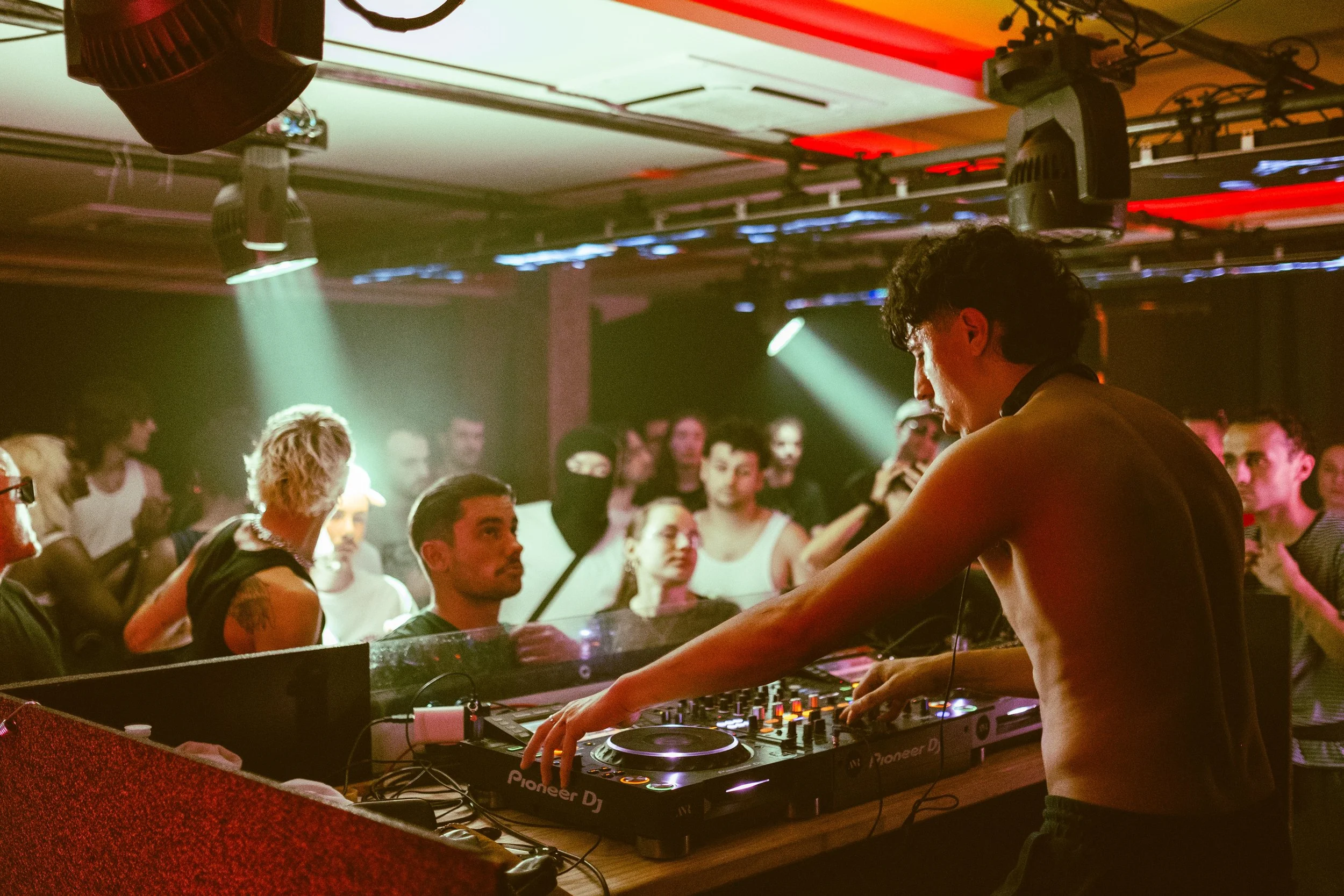 DJ en train de mixer sur scène avec une foule de spectateurs dans un club ou une discothèque, éclairage coloré rouge et vert.