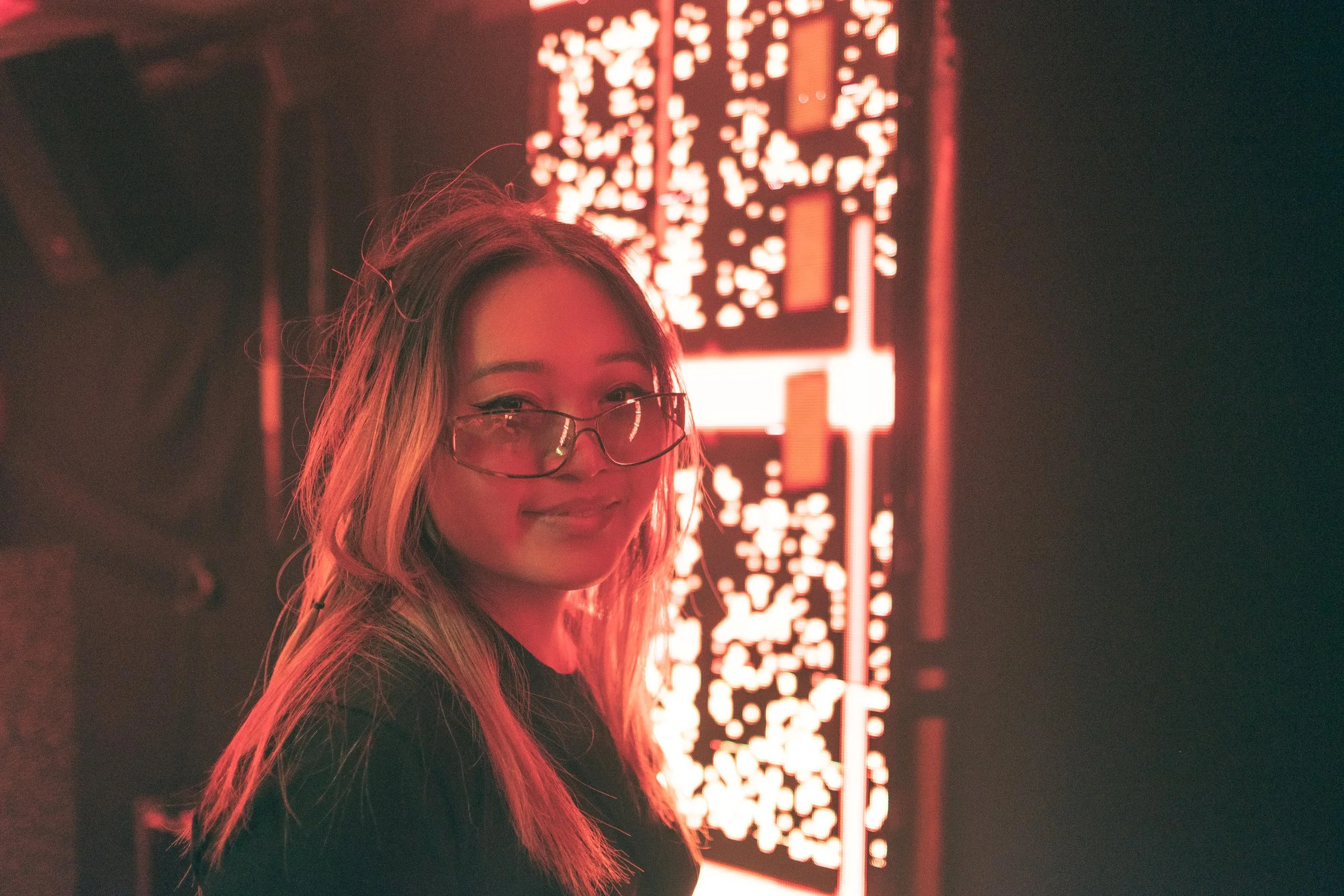 Une femme portant des lunettes, souriante, avec des cheveux longs et blonds, se tient devant une grande installation lumineuse rouge et blanche dans un environnement sombre.