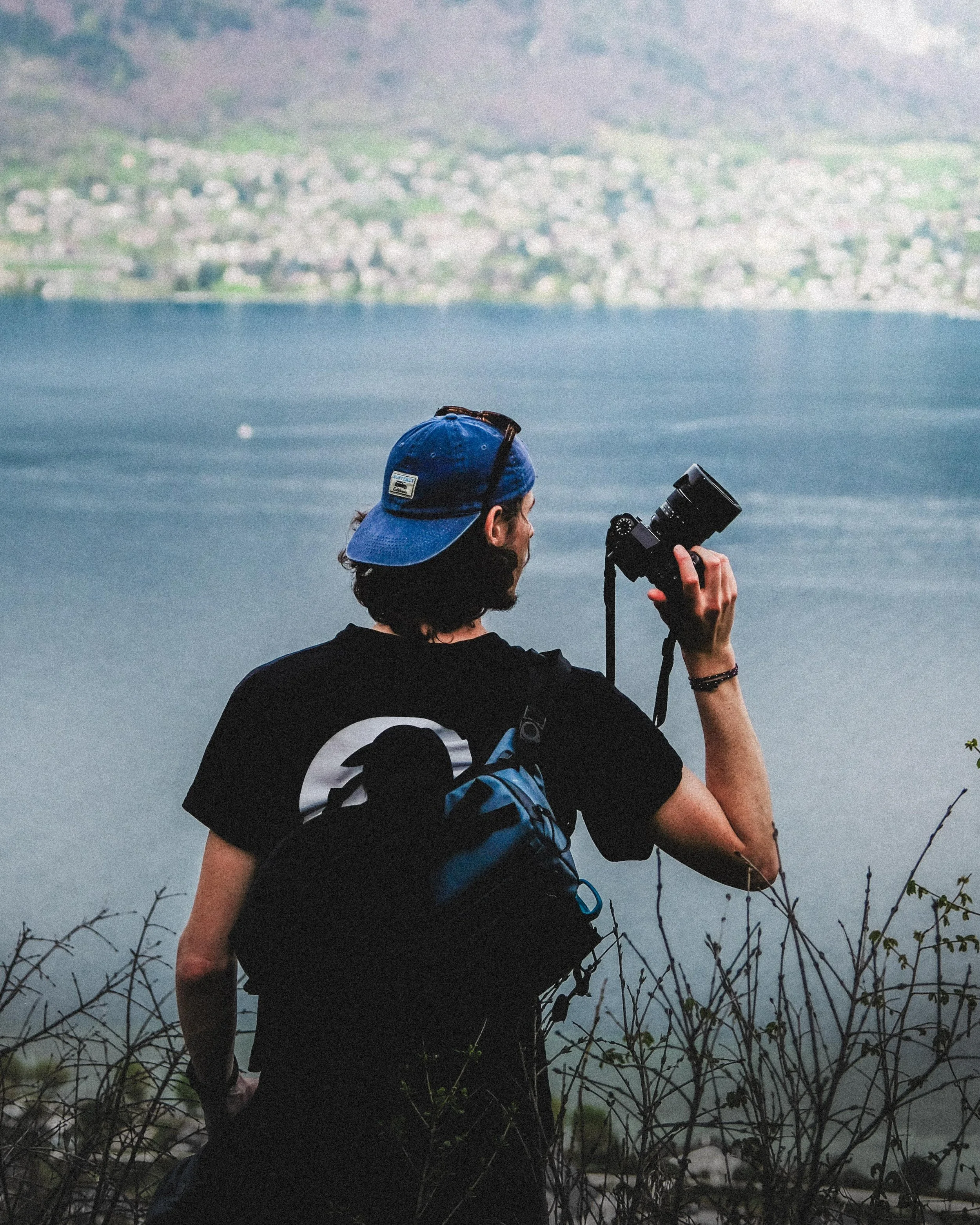 Photographe portant une casquette bleue et tenant un appareil photo devant un paysage côtier brumeux.