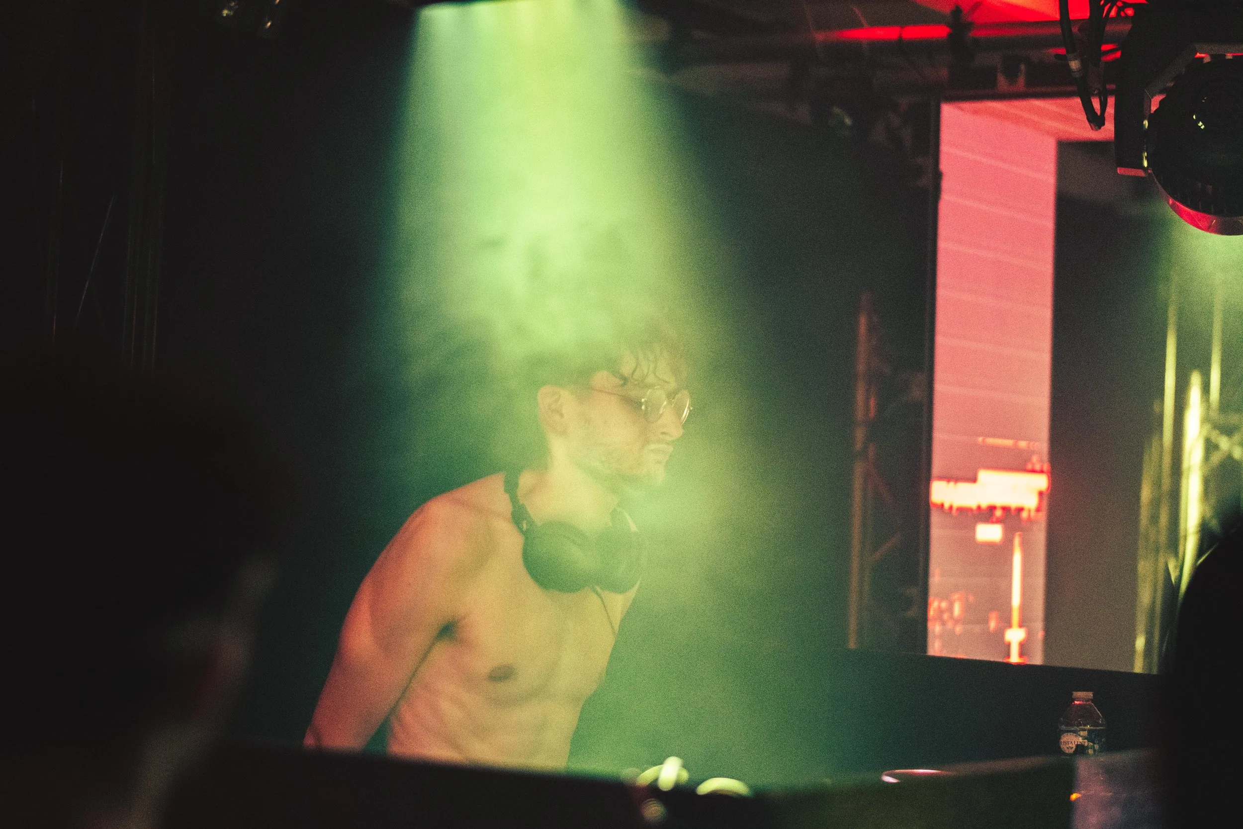 Un DJ avec des écouteurs autour du cou, portant un t-shirt sans manches, jouant dans un club ou lors d’un événement avec un éclairage coloré vert et rouge.