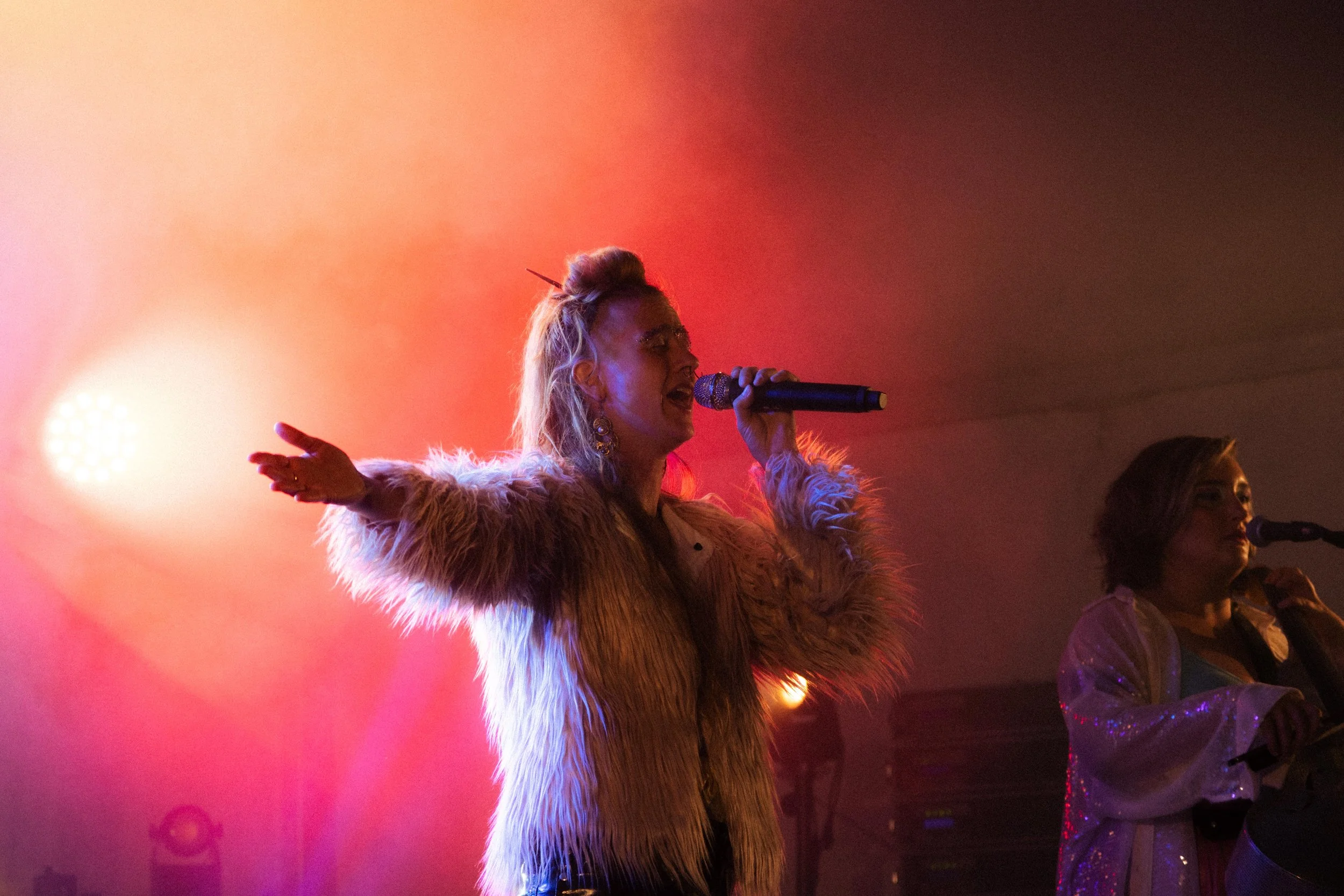 Une femme chante en microphone, portant une veste en fourrure, avec un fond lumineux rouge et une personne derrière elle.