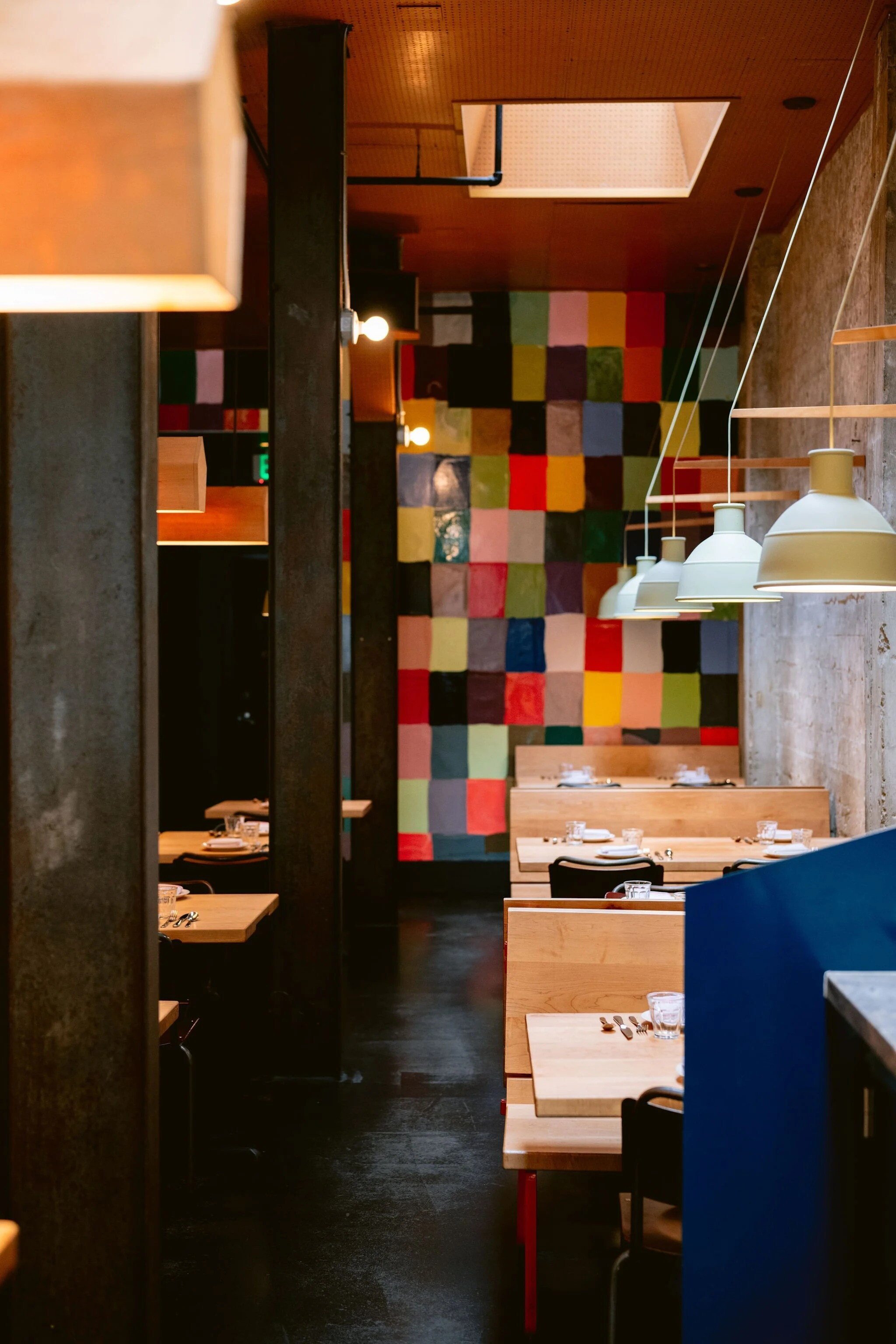 Interior of a modern restaurant with wooden tables set with glasses and silverware, colorful tiled wall as a backdrop, hanging white pendant lights, and a rustic brick wall on the right.
