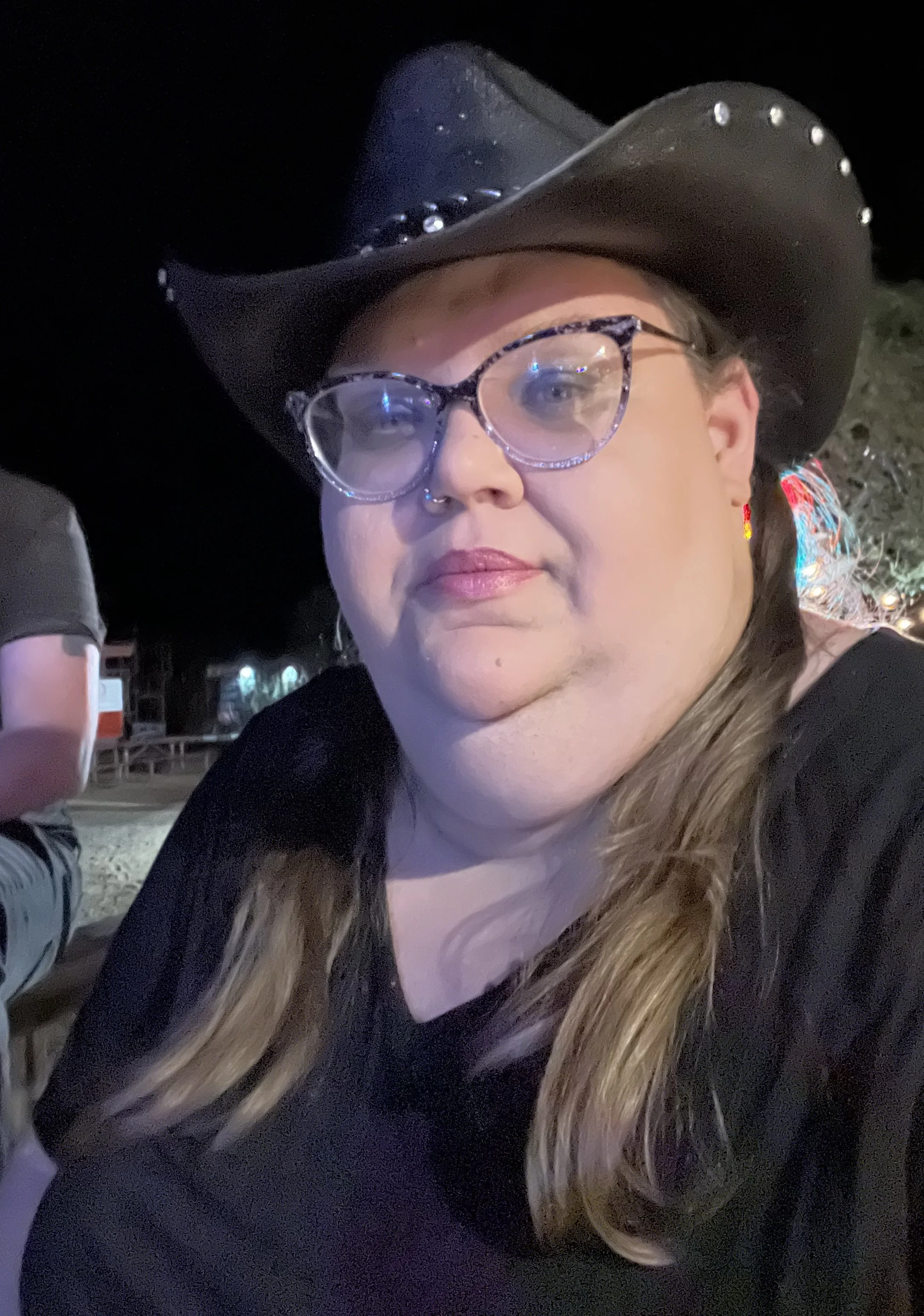 A person with long hair, glasses, and a septum piercing taking a selfie. They are wearing a black top with cut-outs and a silver necklace.