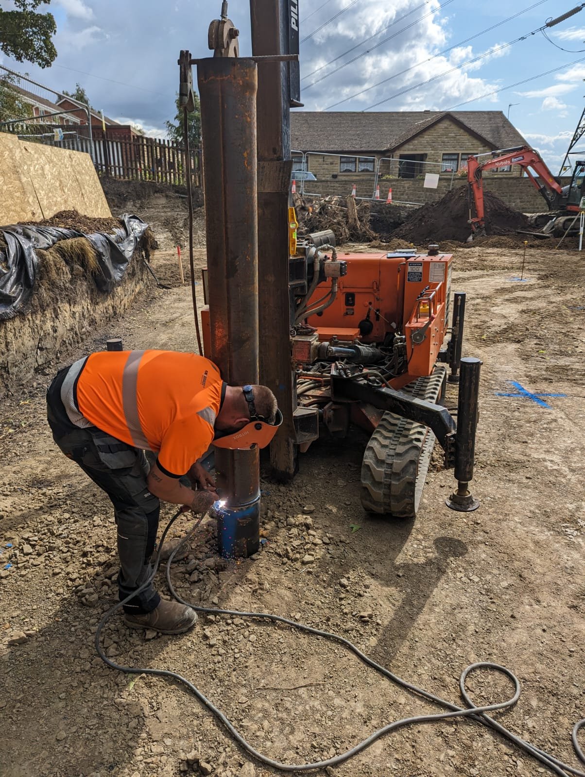 Worker in orange shirt welding a metal post on a construction site with excavation and construction equipment in the background.
