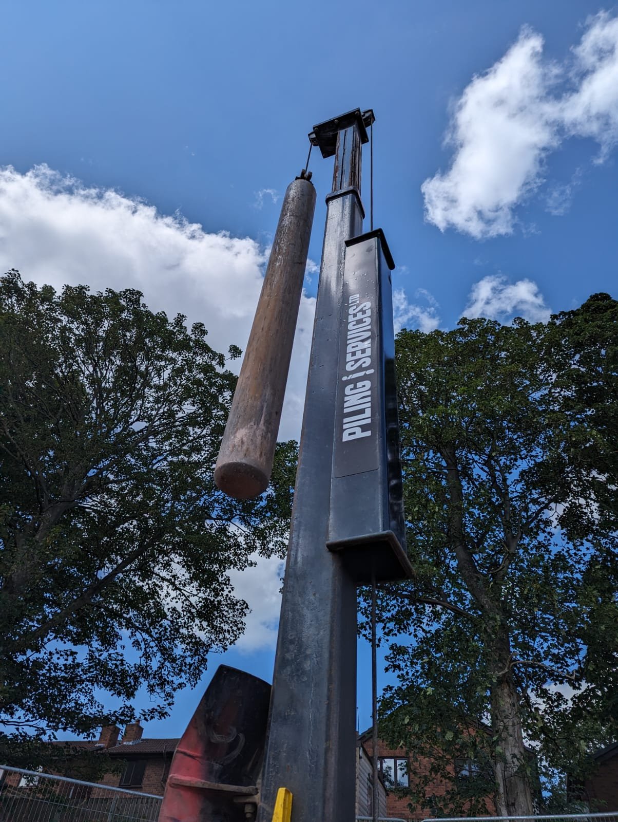 A tall piling machine labeled 'PILING SERVICES' with a large cylindrical wooden pile attached, set against a partly cloudy sky with trees and residential buildings in the background.