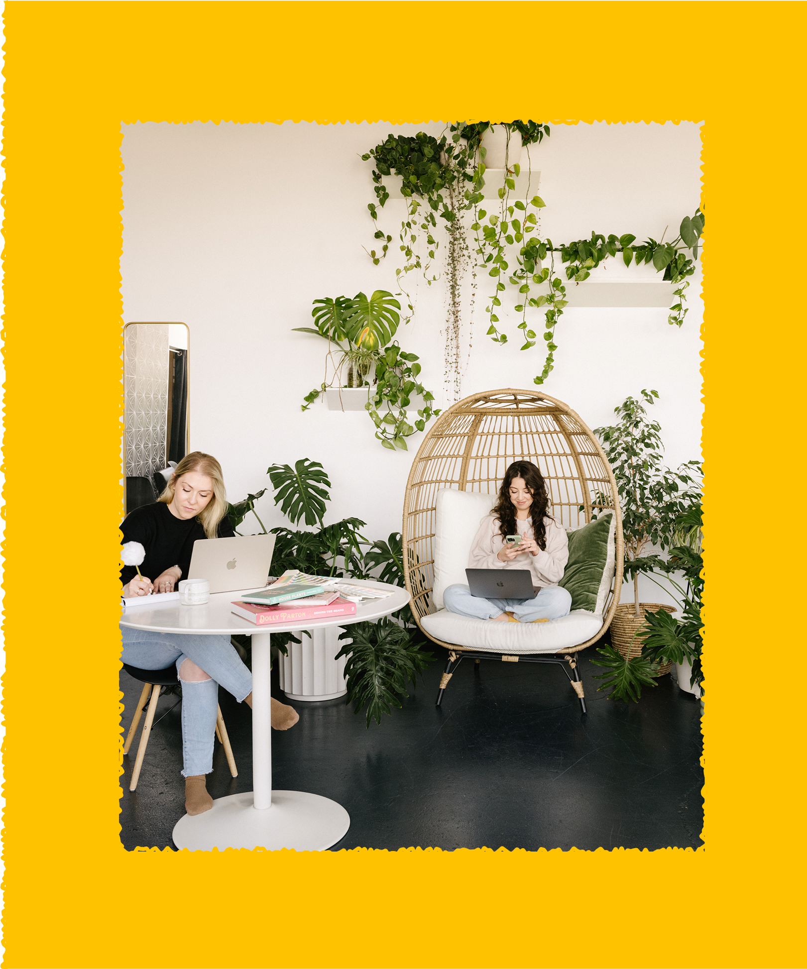 Two women working in a modern, plant-filled room. One seated at a round white table with a laptop and notebooks; the other sitting in a wicker hanging chair with a laptop, using her phone. The room has white walls, green plants, and a large mirror.